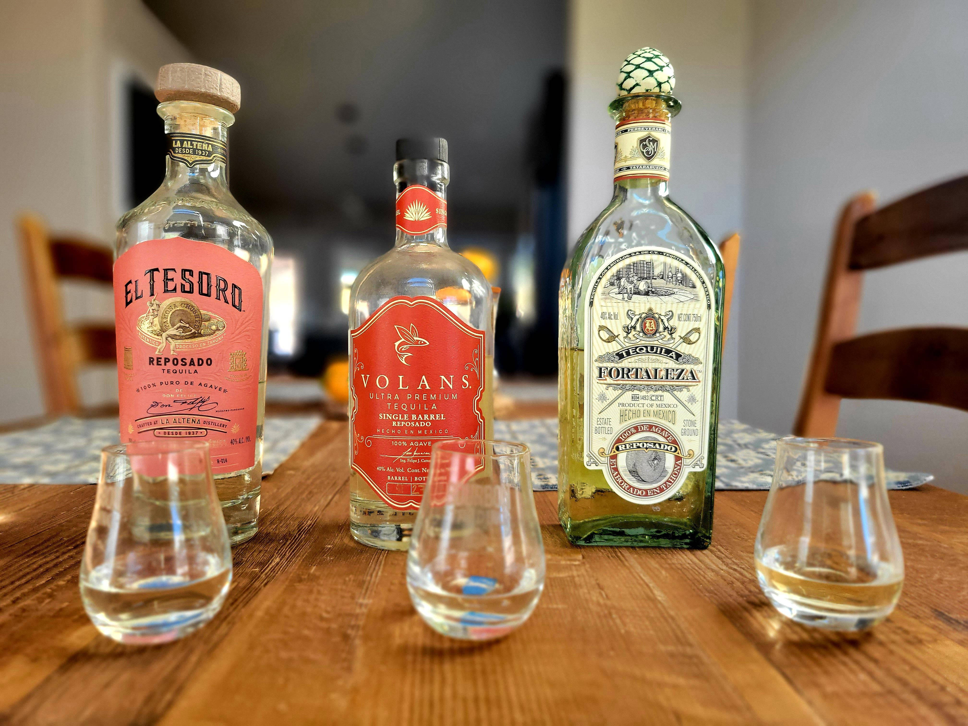 alcohol 桌布 壁紙 — 三種不同的龍舌蘭酒瓶配品酒杯，擺放在一張鄉村風格的木桌上，室內燈光溫暖。