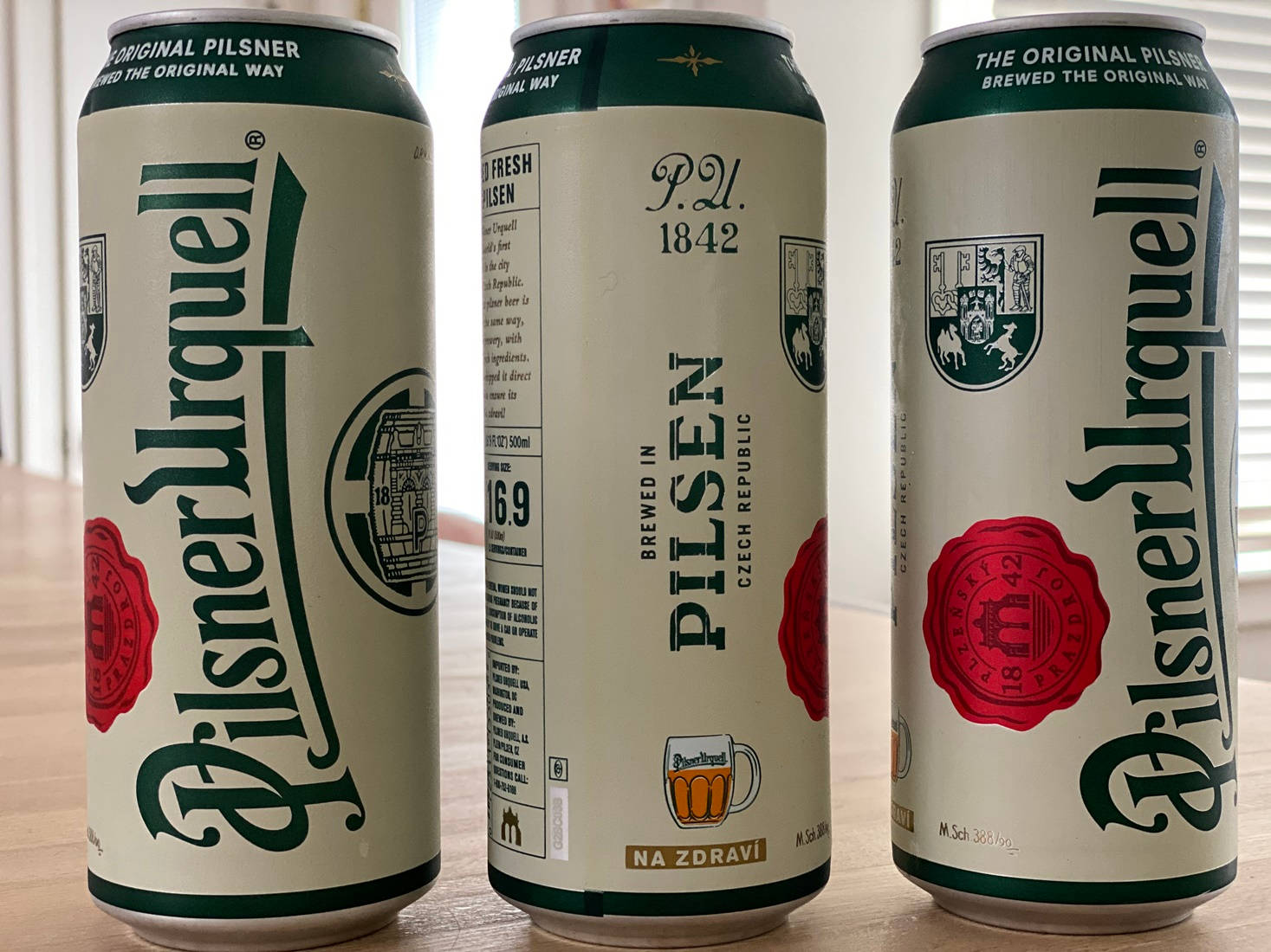 Beer 桌布 壁紙 — 三罐奶油色、綠色和紅色的Pilsner Urquell啤酒罐並排放在窗邊的木桌上。