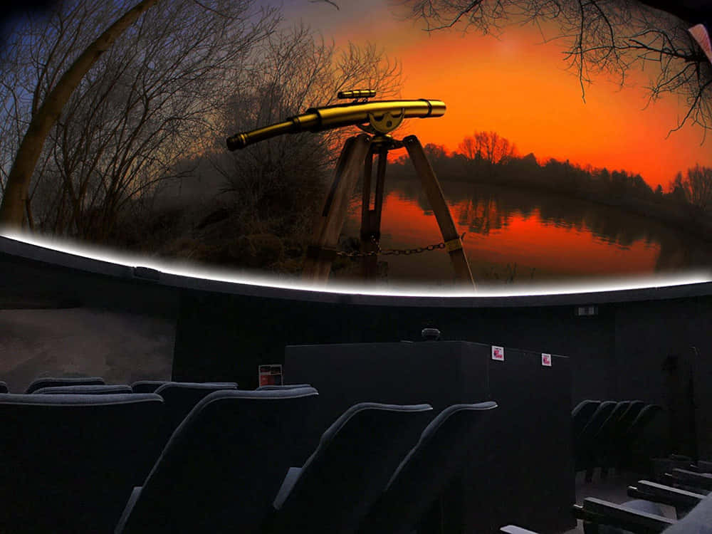 Planetarium 桌布 壁紙 — 三腳架上的金色望遠鏡指向平靜的湖面上反射出的火橙色日落，湖邊環繞著光禿禿的樹木。