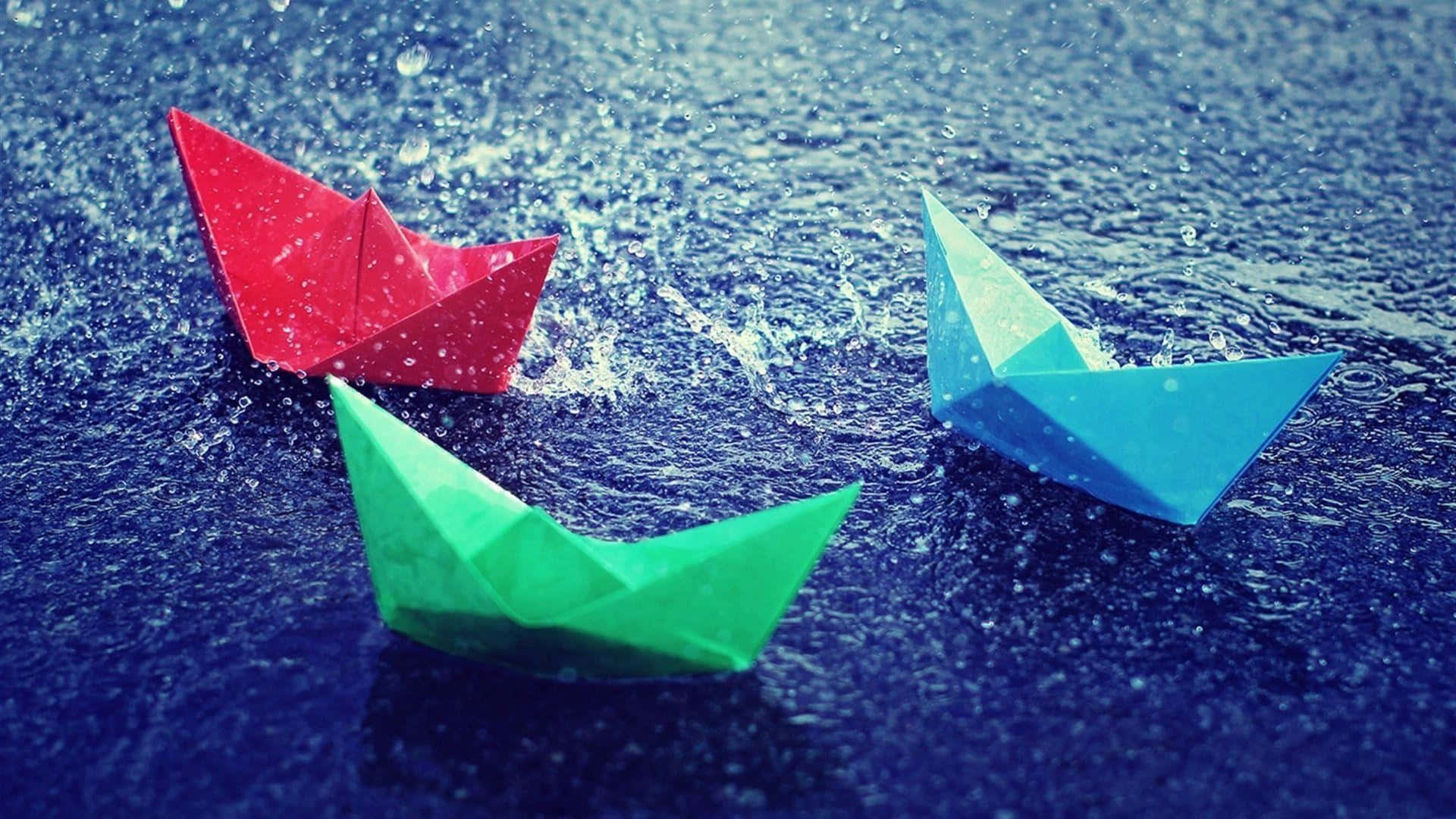 Rain Drops 桌布 壁紙 — 三艘色彩繽紛的摺紙船，紅色、綠色和藍色，在帶有紋理的藍色水面上航行，水花四濺。