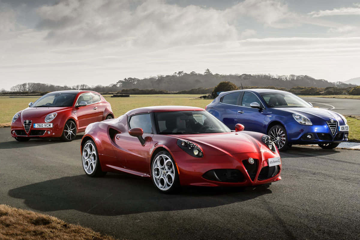 愛快羅密歐 桌布 壁紙 — 三輛 Alfa Romeo 汽車，紅色、藍色和紅色跑車停在瀝青路上，後面有草地和陰天。