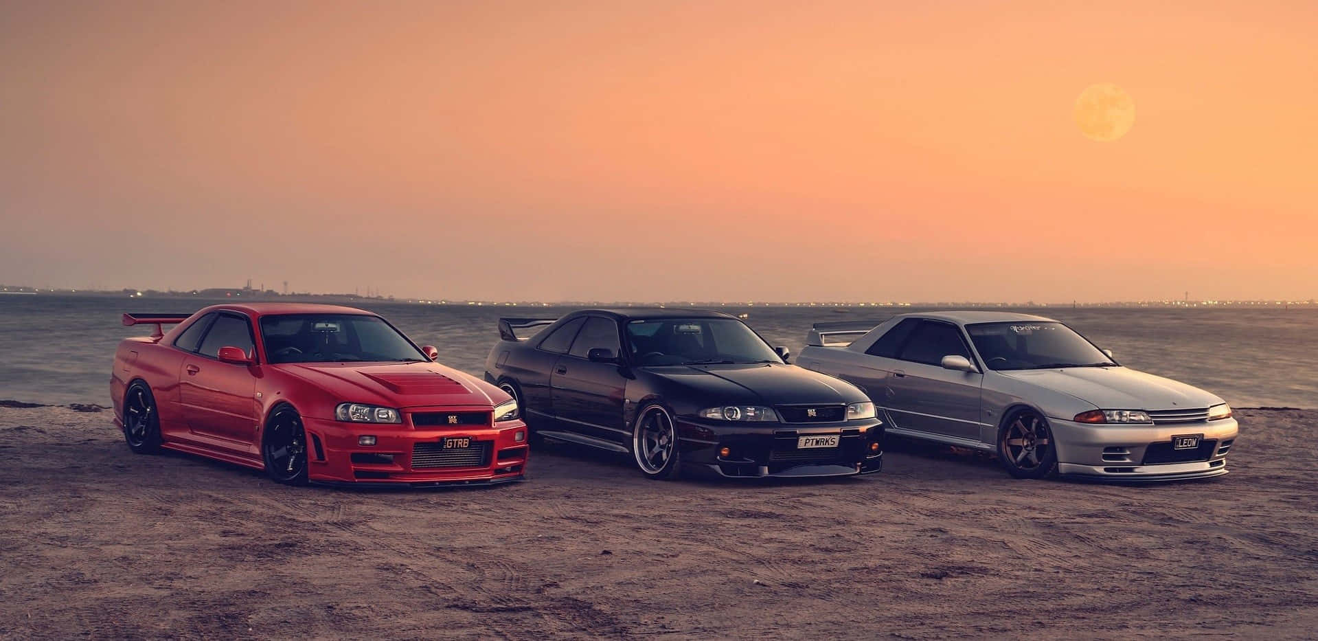 R33 GT-R 桌布 壁紙 — 三輛改裝日產Skyline GT-R停泊在日落時分的沙灘上，上方有一輪發光的月亮。