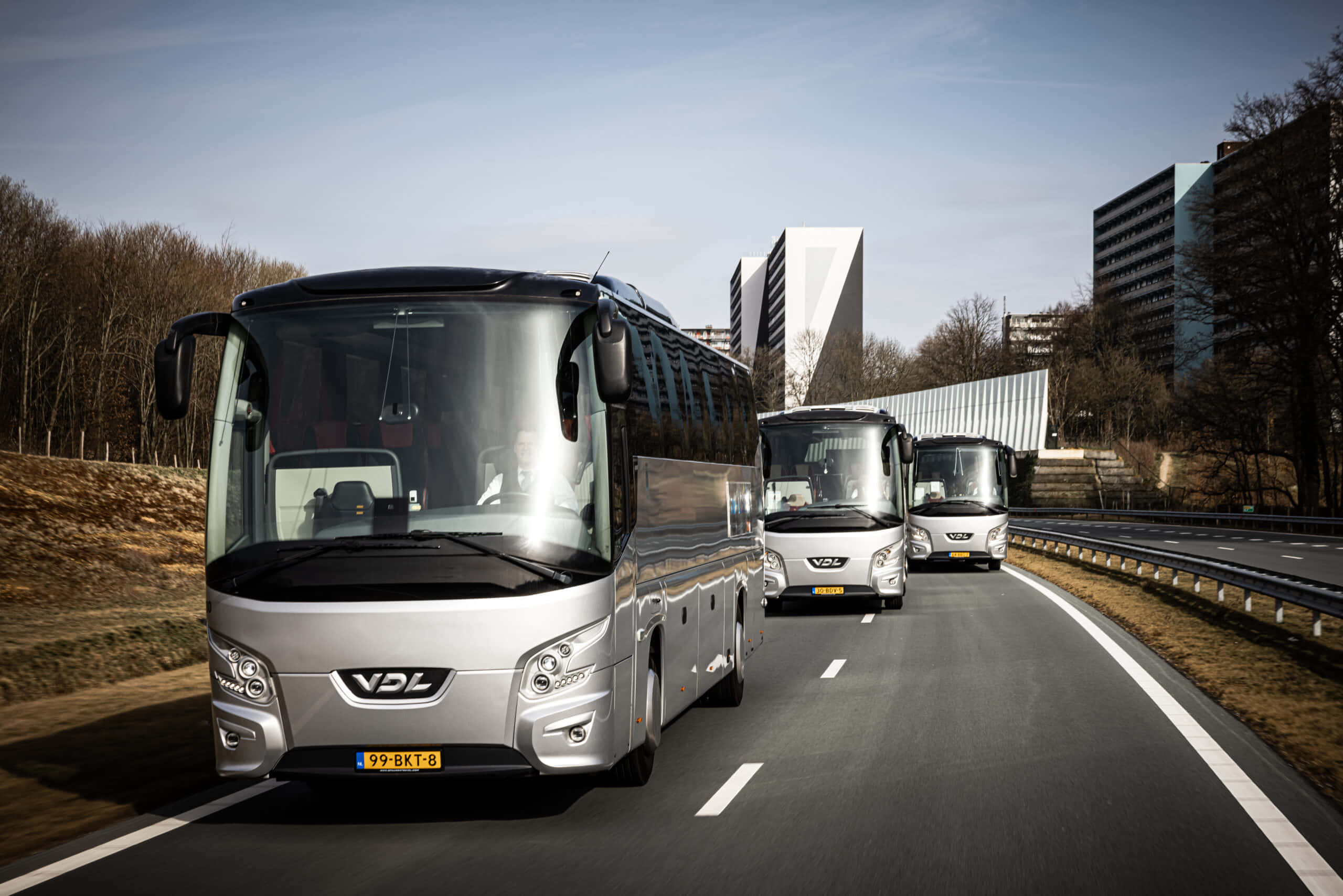 Bus 桌布 壁紙 — 三輛時尚的銀色VDL客車在陽光明媚的高速公路上以隊形行駛，旁邊是現代建築和光禿禿的樹木。