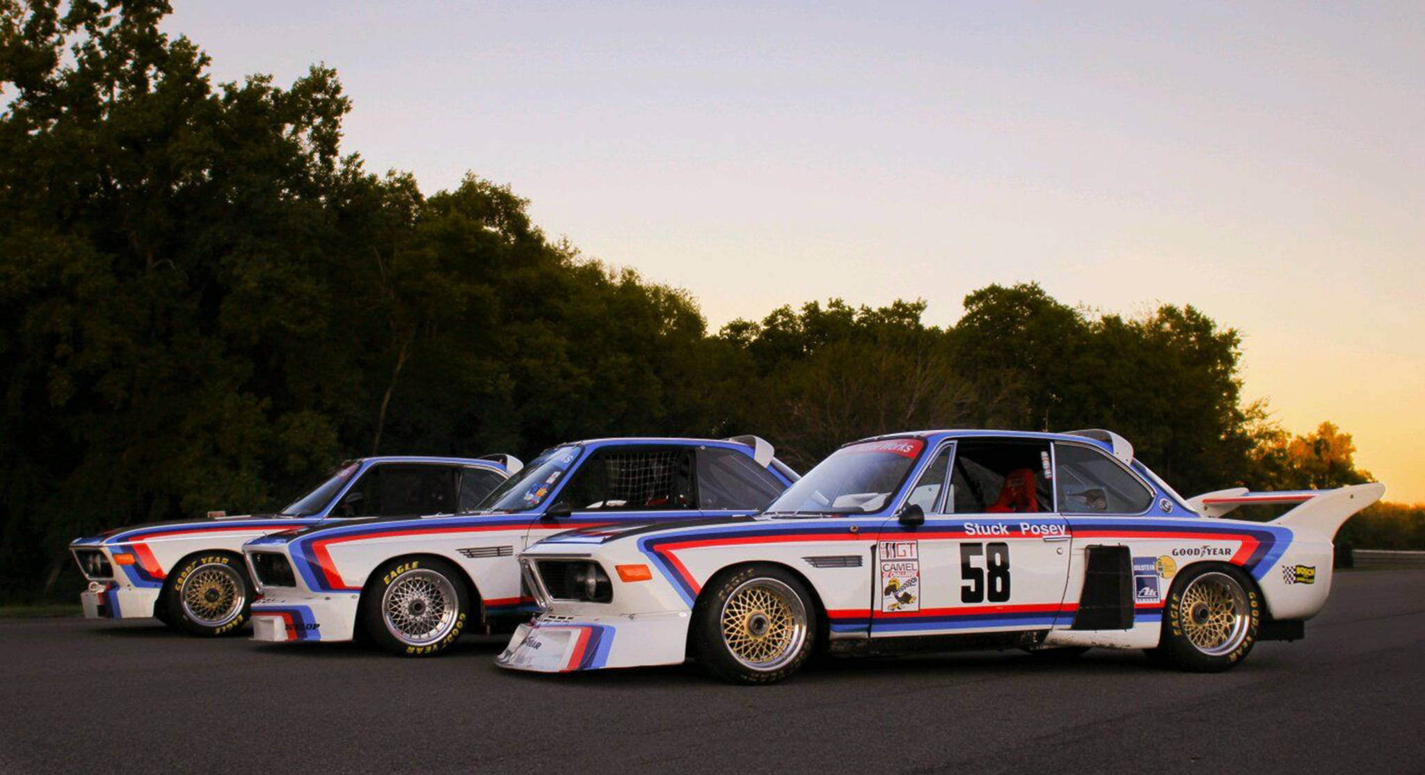 classic bmw 桌布 壁紙 — 三輛標誌性寶馬賽車，採用藍、紅、白塗裝，在黃金時刻停泊在瀝青路上，後面有樹木。