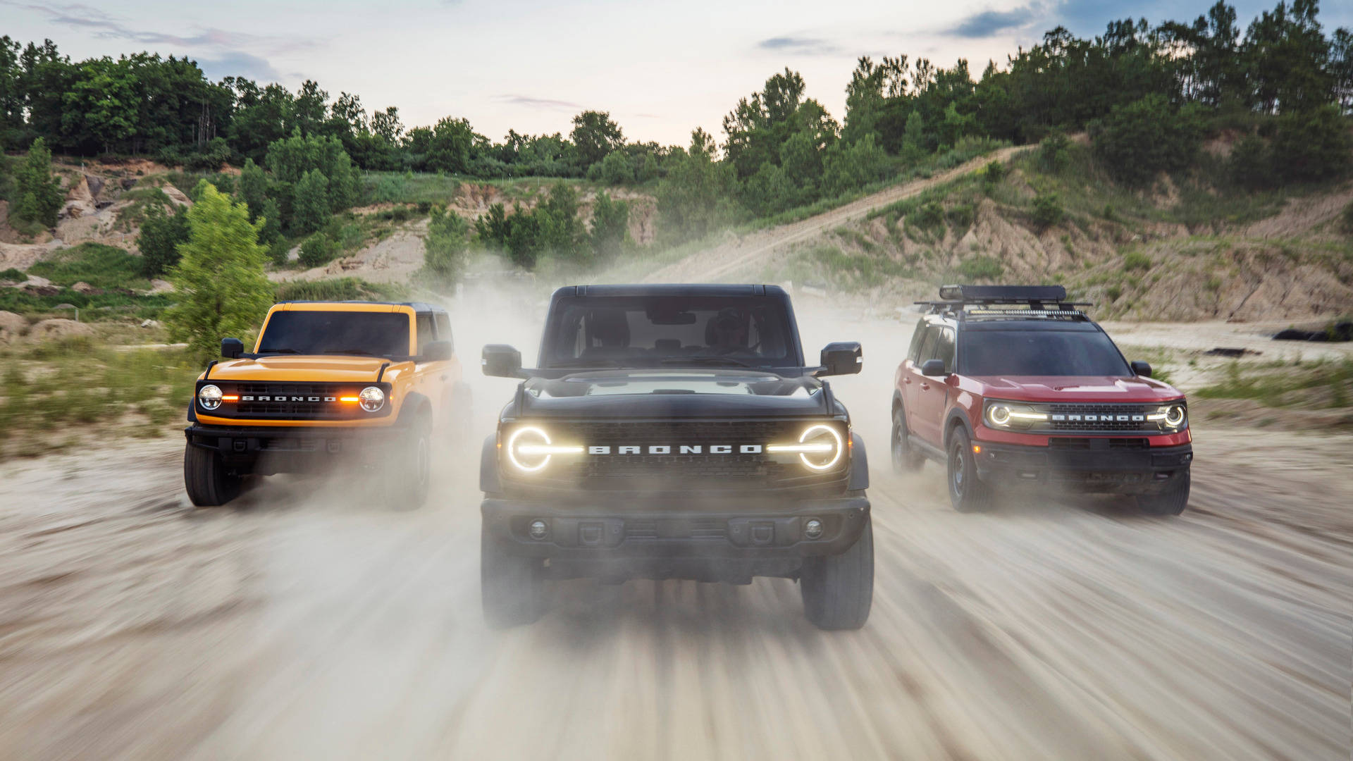 Ford Bronco 桌布 壁紙 — 三輛橙色、黑色和紅色的福特 Bronco 在塵土飛揚的小路上咆哮，揚起滾滾塵土，背景是樹木繁茂的山坡。