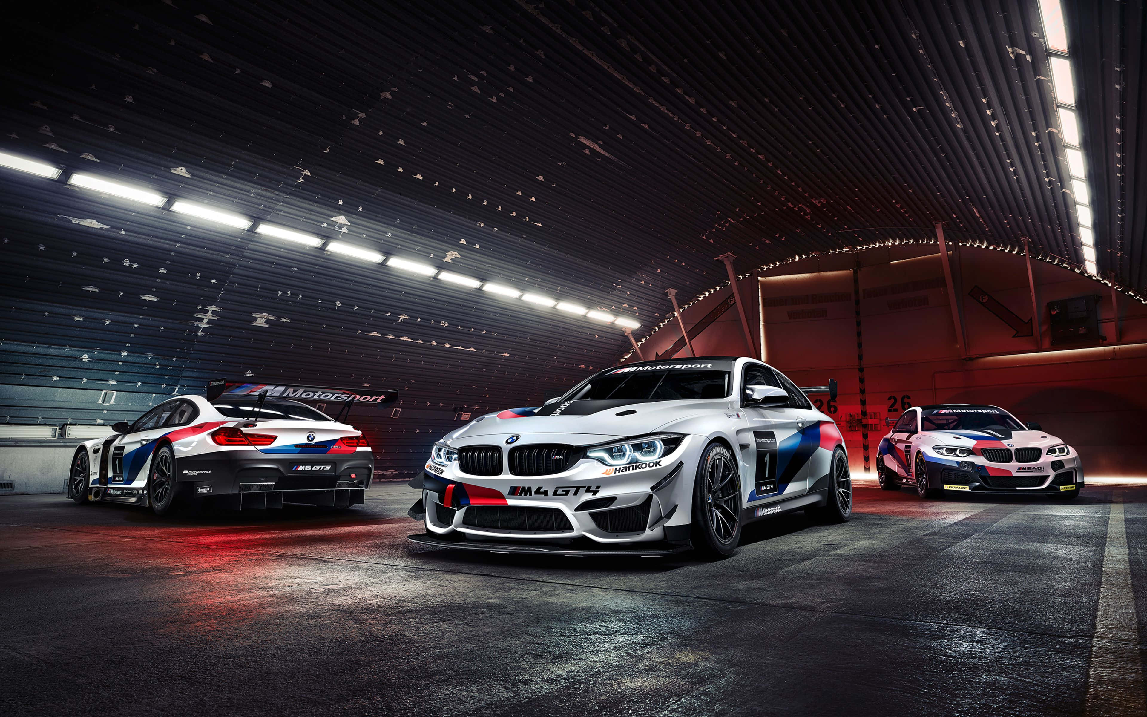 BMW 動圖 桌布 壁紙 — 三輛白色、藍色和紅色塗裝的BMW M4 GT4賽車停放在時尚的現代化車庫中，上方有明亮的頂燈。