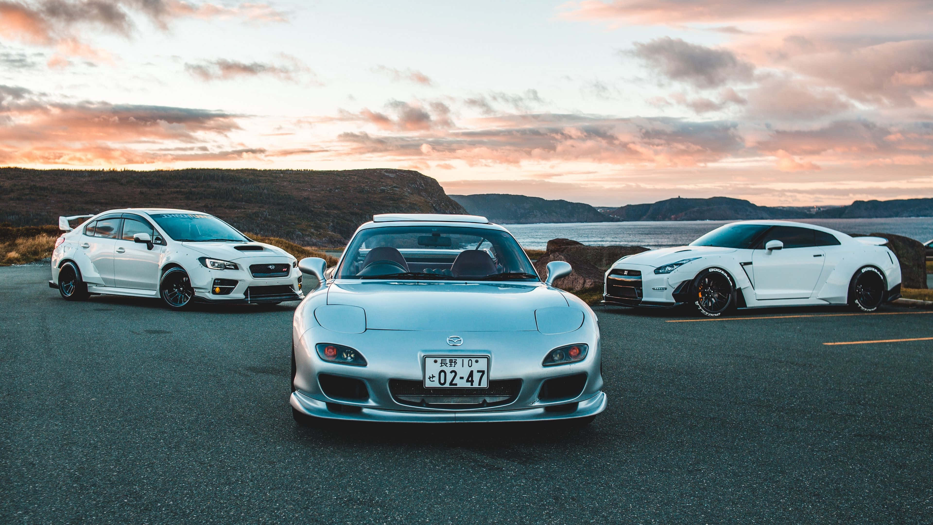 Mazda 桌布 壁紙 — 三輛白色高性能汽車停泊在日落時分的沿海道路上，上方有粉紅色和橙色的雲彩。