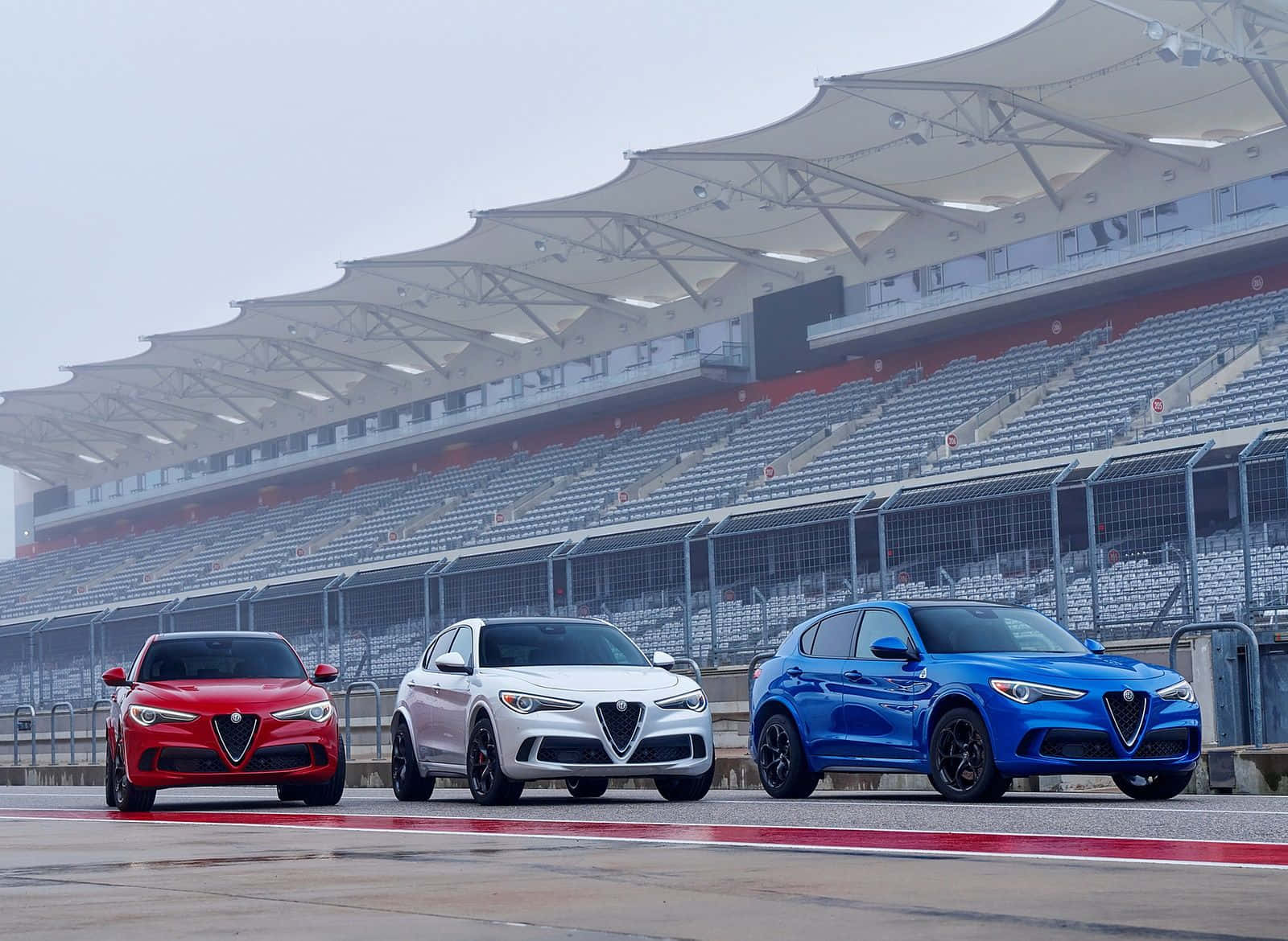 Alfa Romeo Stelvio 桌布 壁紙 — 三輛紅色、白色和藍色的愛快羅密歐SUV在陰天下一字排開在賽車道上，後面是空無一人的看台。
