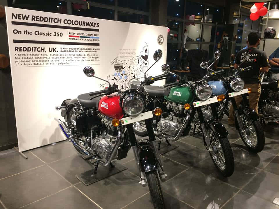 New 桌布 壁紙 — 三輛紅色、綠色和藍色的 Royal Enfield Classic 350 摩托車在室內明亮燈光下展示，靠近資訊牌。