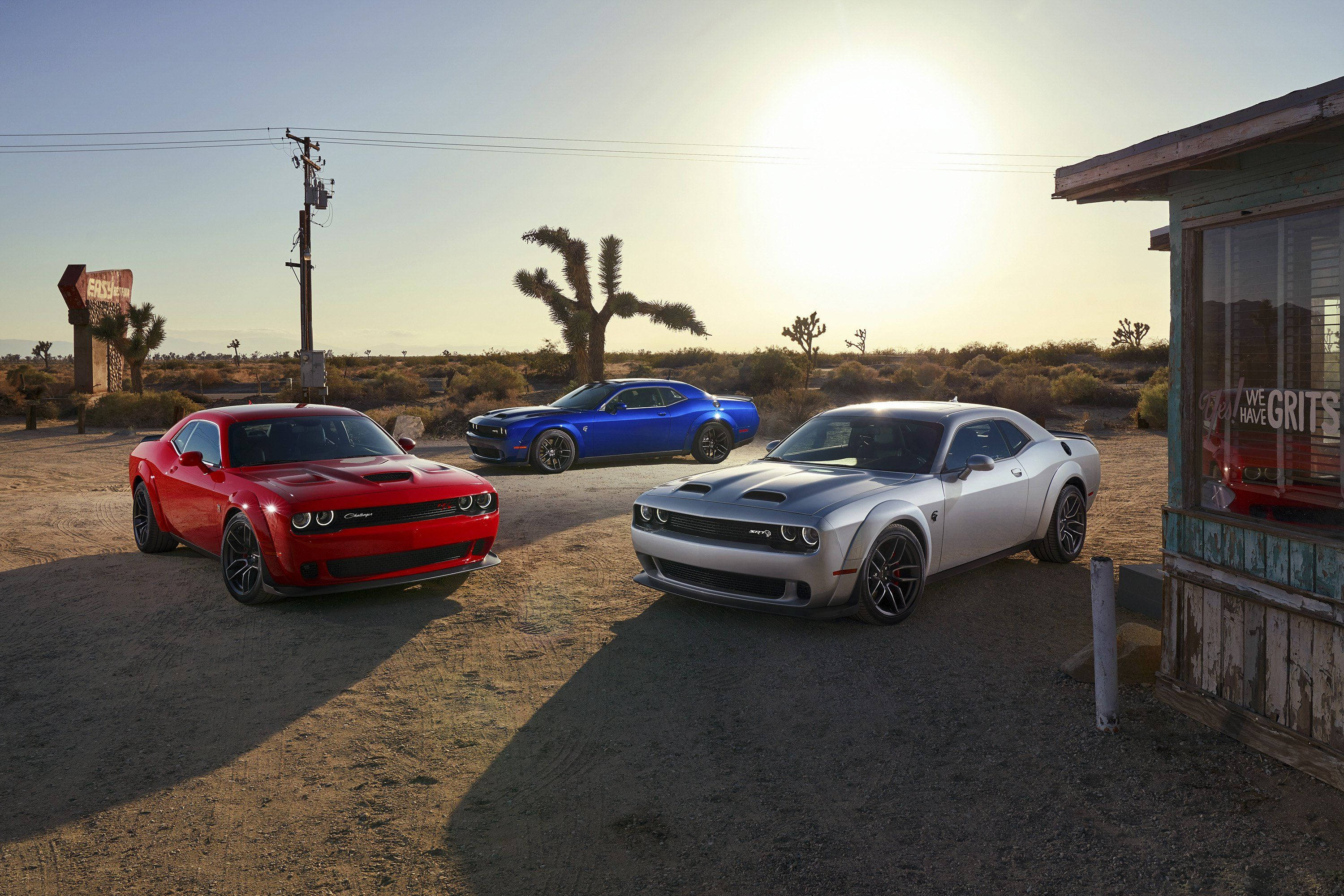 Challenger 桌布 壁紙 — 三輛肌肉車（紅色、藍色和銀色）停在塵土飛揚的沙漠路邊，正值黃金時刻。