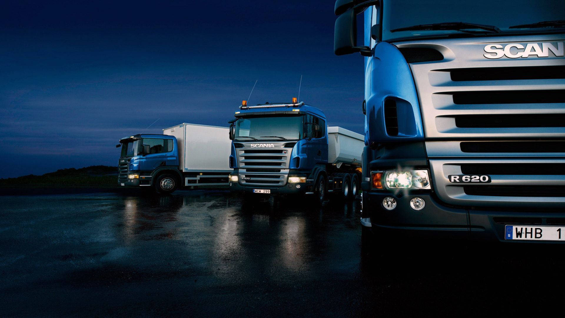Trucks 桌布 壁紙 — 三輛藍色 Scania 貨車停放在濕漉漉的瀝青場地上，深藍色的暮色天空下，車頭燈反射在地面上。