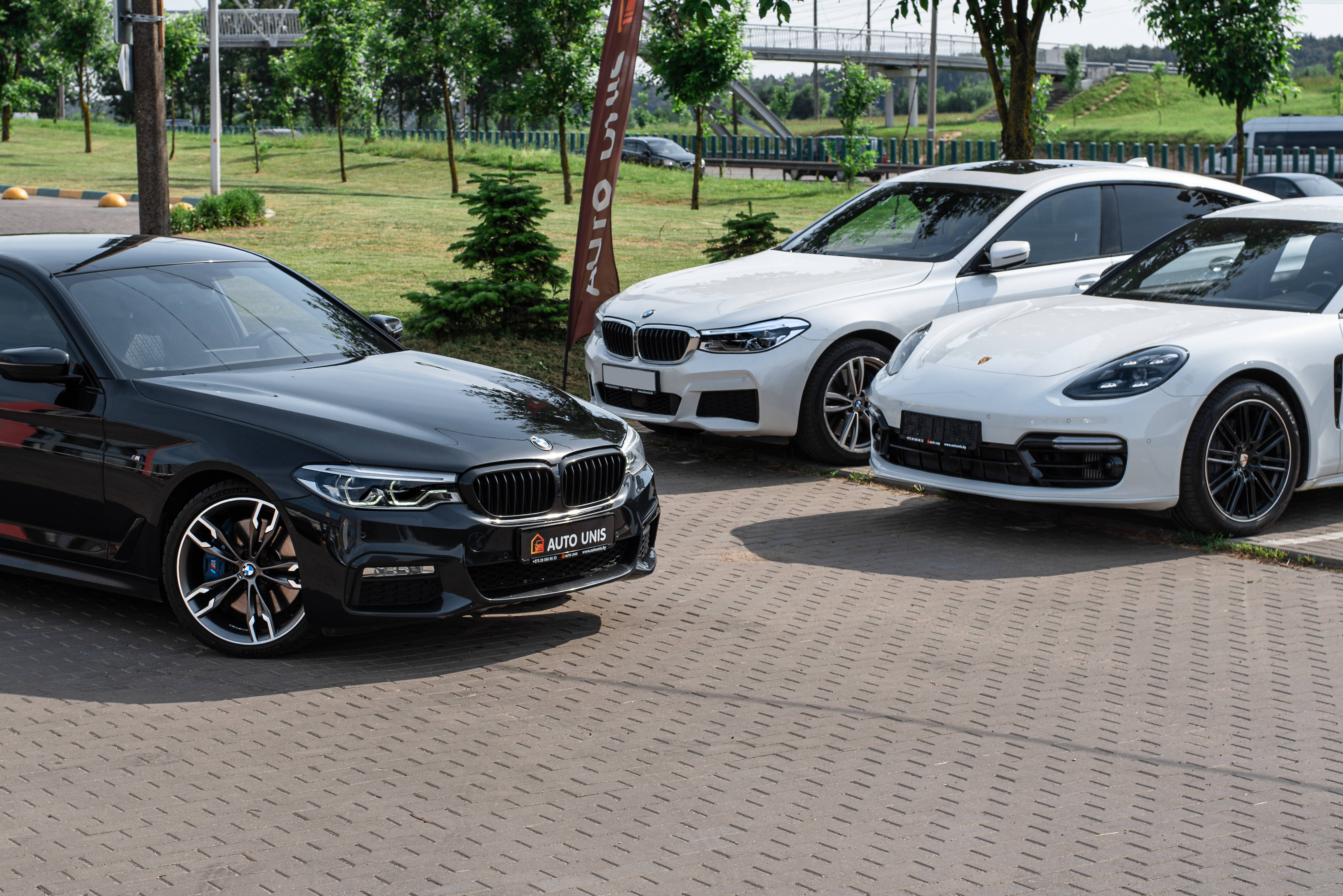 m 桌布 壁紙 — 三輛豪華車 黑色BMW轎車 白色BMW轎車 白色保時捷Panamera 停放在鋪砌的停車場 背景有綠樹和橋樑