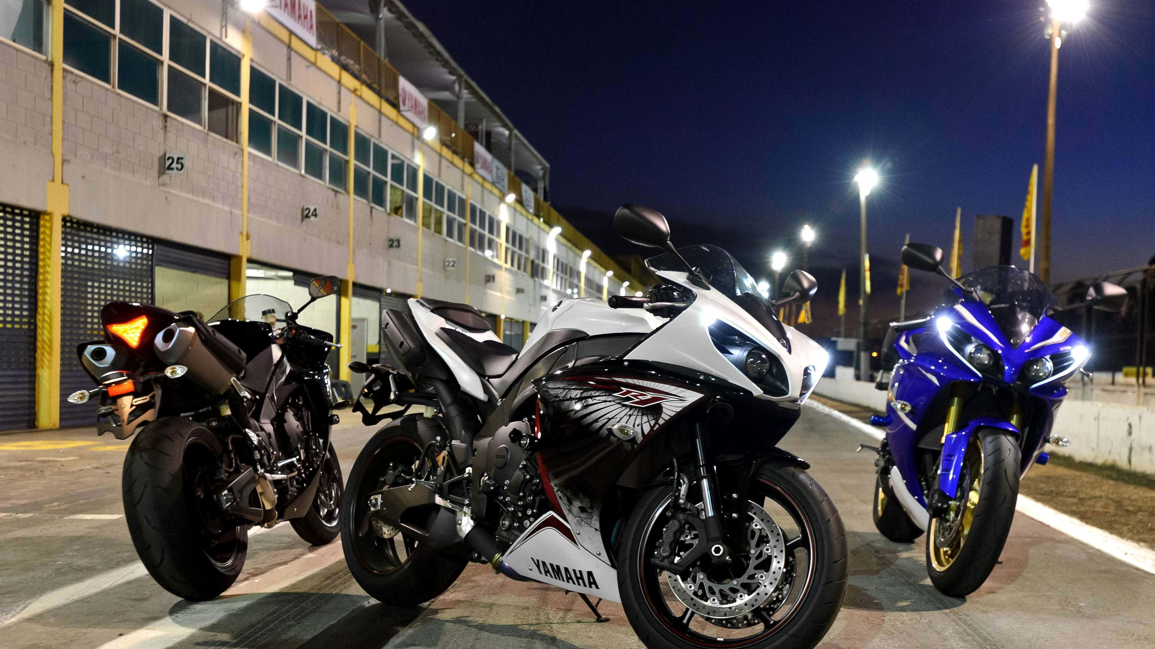 4K Bikes 桌布 壁紙 — 三輛高性能Yamaha跑車在夜間停放在賽車設施外，在明亮的泛光燈下閃閃發光。