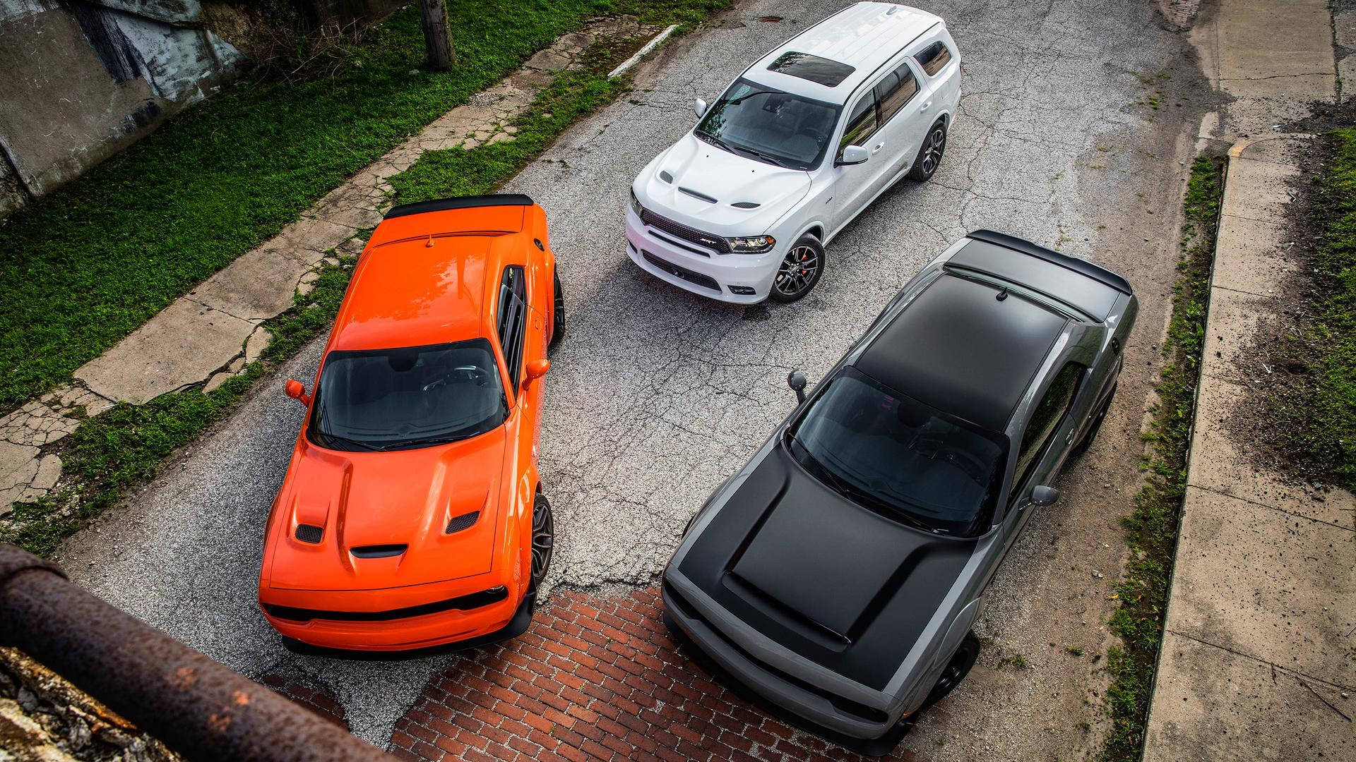 Dodge 桌布 壁紙 — 三輛高性能的Dodge Challengers，分別為橙色、白色和消光黑色，停放在一條有草叢邊緣的裂縫柏油路上。
