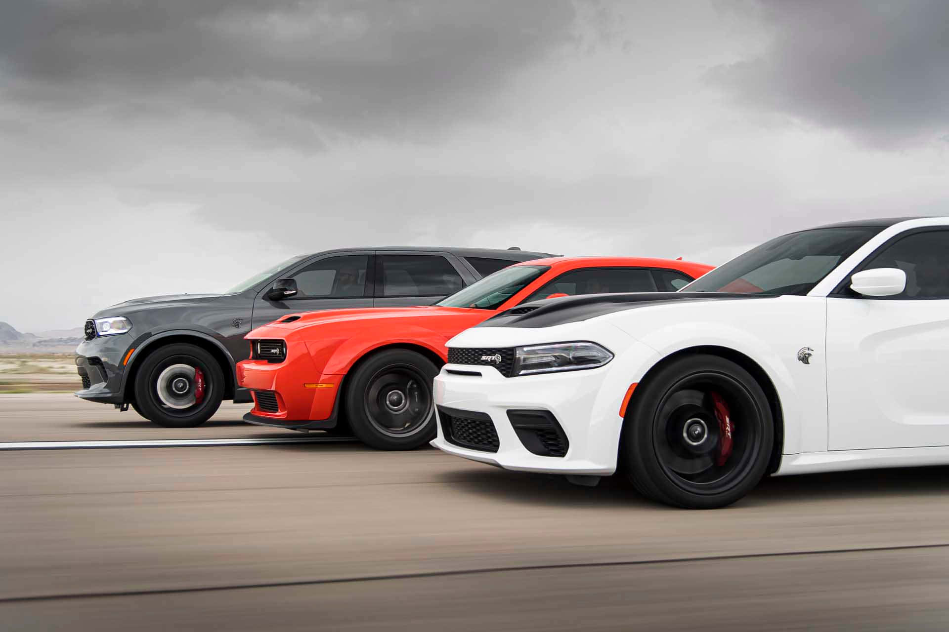 Dodge 桌布 壁紙 — 三輛高性能肌肉車（灰色、紅色和白色）在暴風雨的天空下沿沙漠高速公路飛馳。