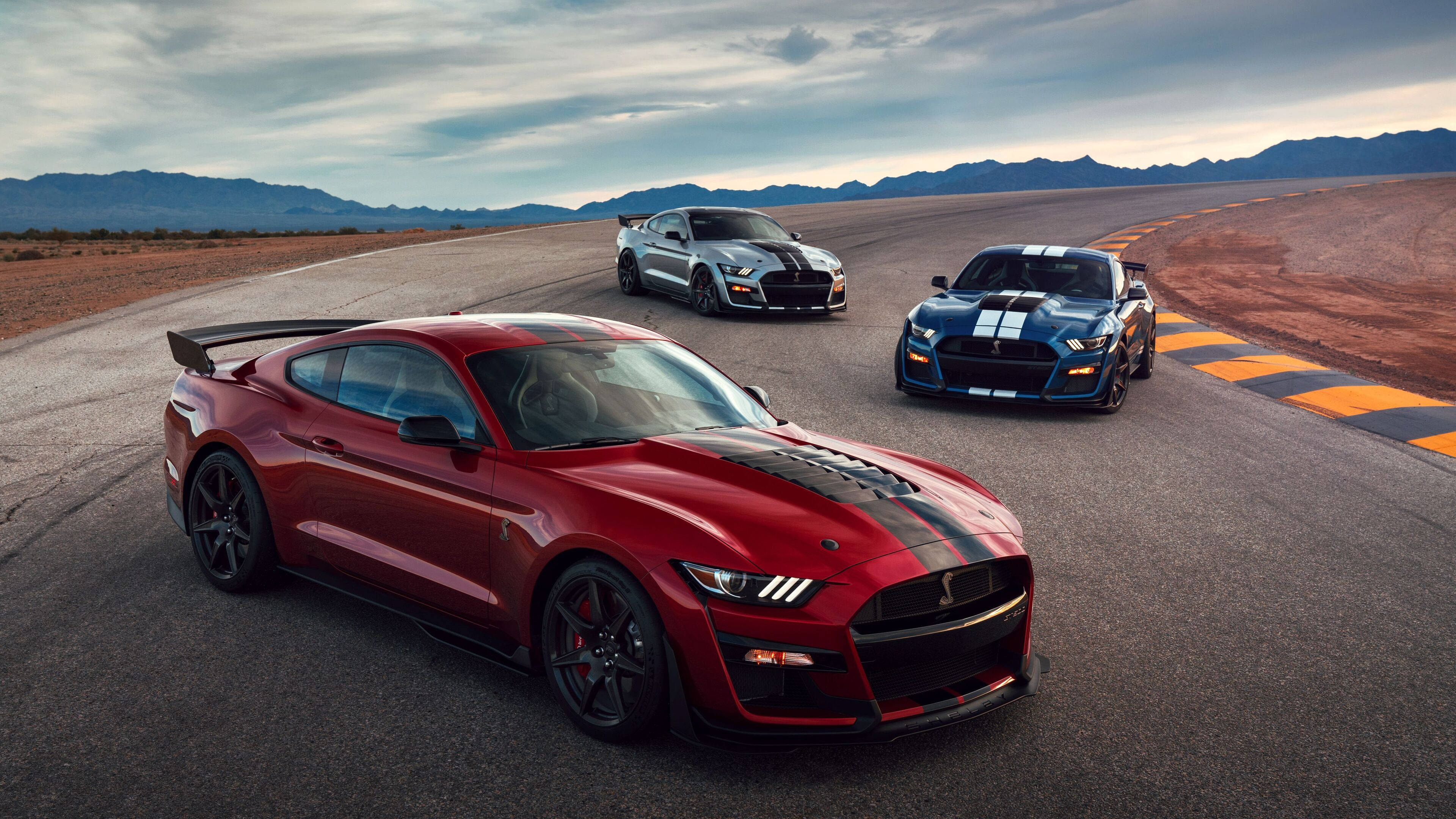 mustang HD 桌布 壁紙 — 三輛高性能野馬在沙漠賽道上奔馳，背景是山脈和陰天。