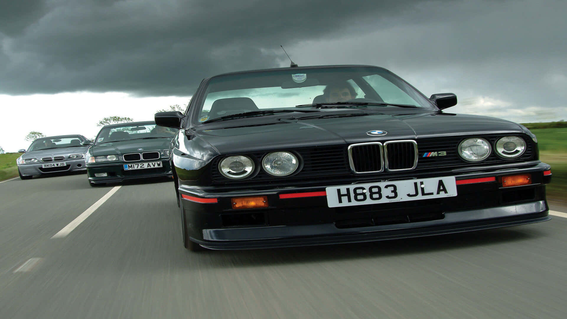 Bmw M Series 桌布 壁紙 — 三輛黑色BMW M3在暴風雨的天空下，以模糊的動態在濕滑的道路上競速。