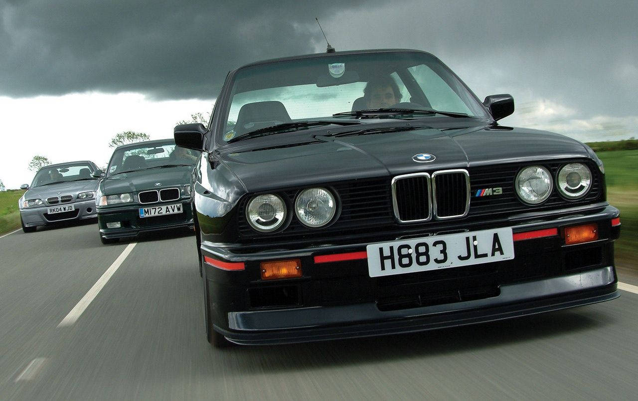 m 桌布 壁紙 — 三輛黑色BMW M3跑車在暴風雨灰暗的天空下，在潮濕的道路上奔馳，呈現模糊的動態效果。