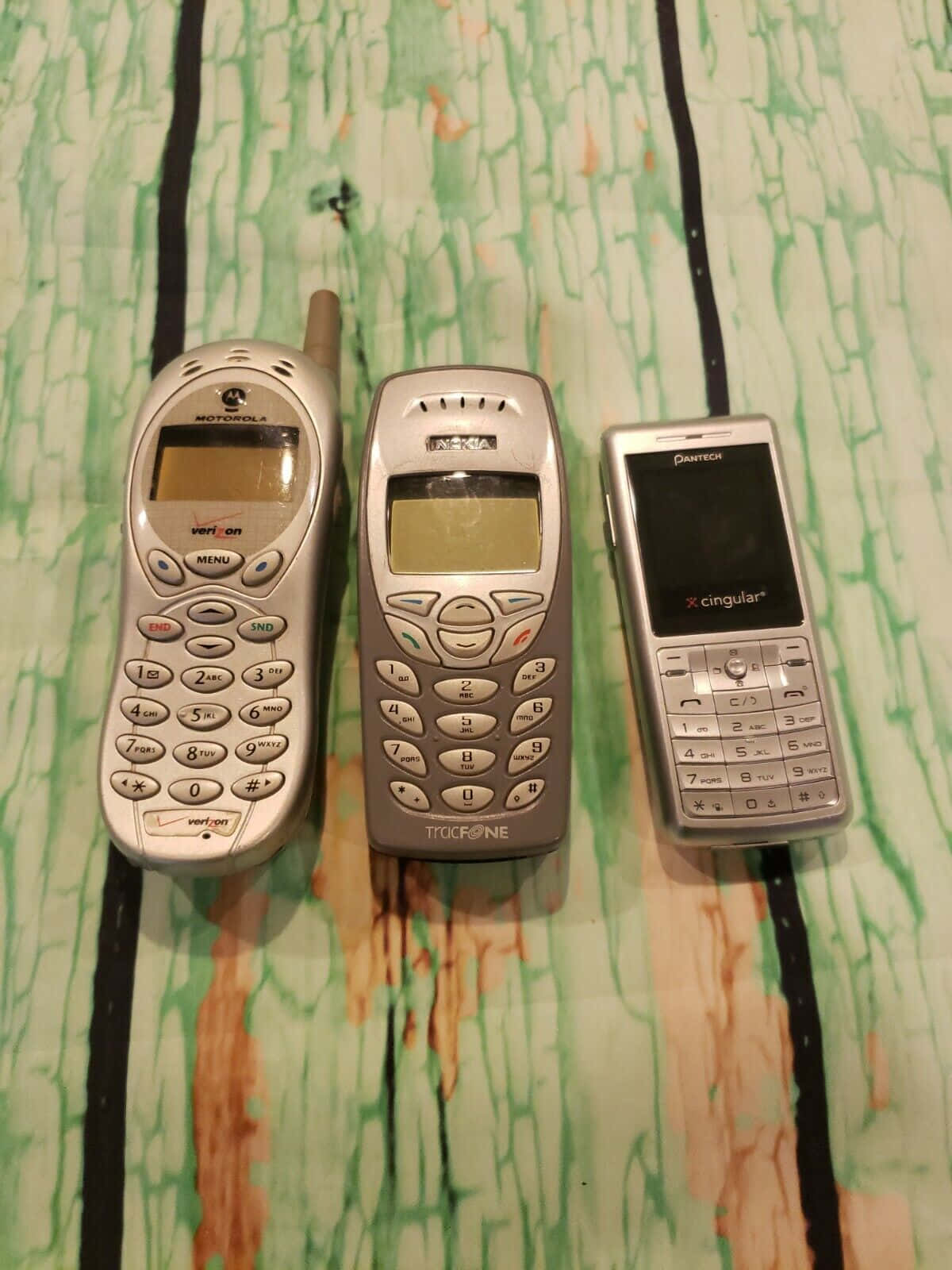 手機 桌布 壁紙 — 三部復古手機 Motorola Nokia 和 Pantech 並排放置在綠色和棕色圖案表面上。