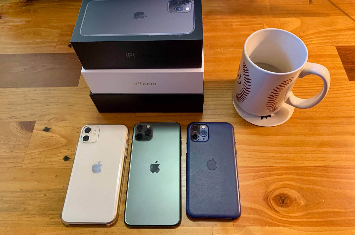 iPhone 11 桌布 壁紙 — 三部白色、綠色和藍色的iPhone放在木桌上，旁邊有它們的盒子和一個棒球杯。