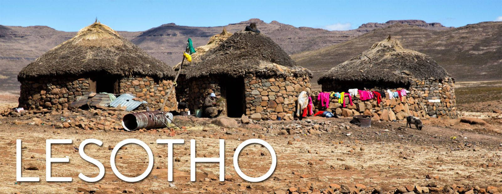 Lesotho 桌布 壁紙 — 三間石頭茅草屋頂的小屋矗立在乾燥的賴索托山谷中，藍天白雲下，附近掛著色彩繽紛的衣物。