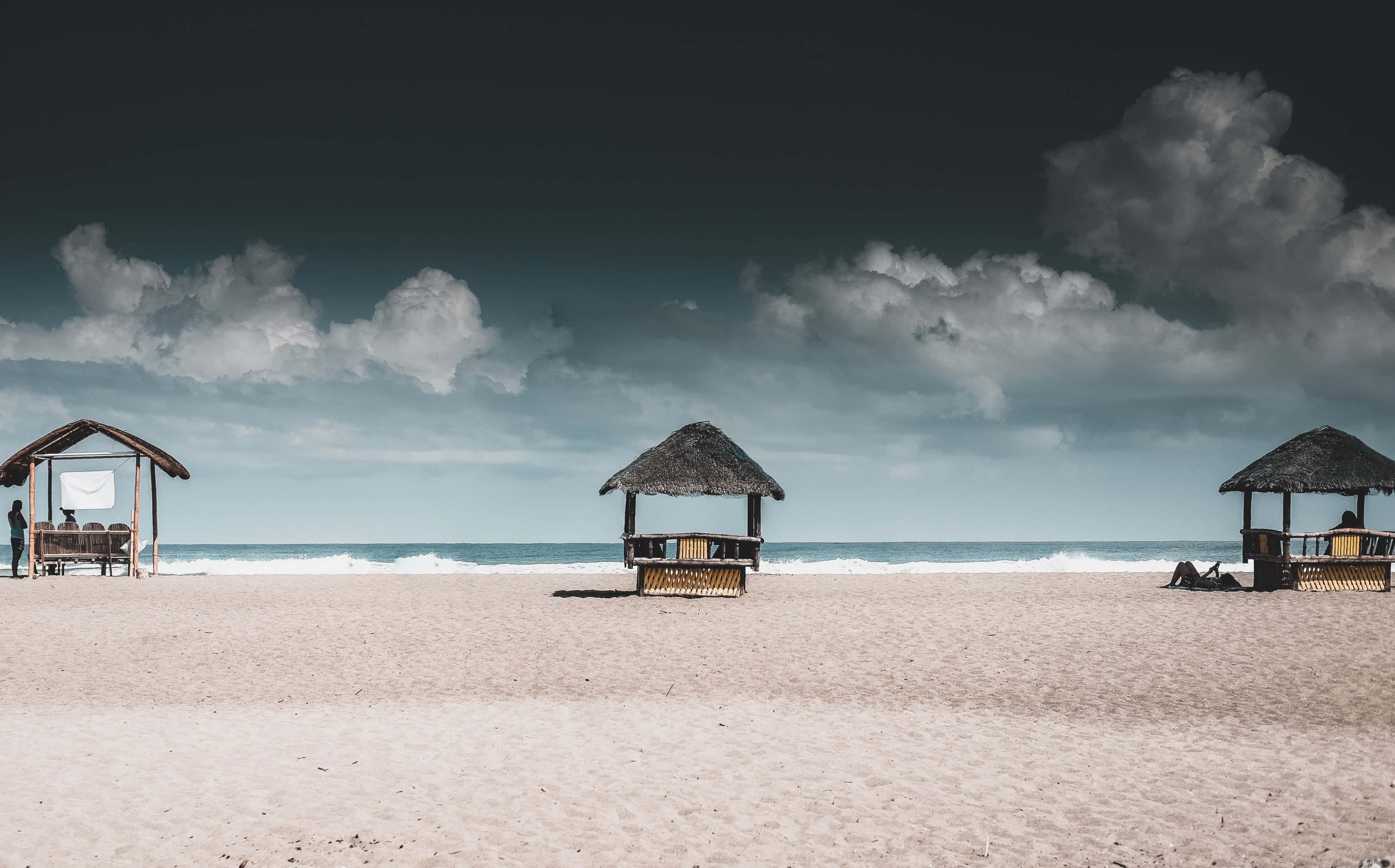 Beach Huts 桌布 壁紙 — 三間茅草屋頂的海灘小屋坐落在寬闊的沙灘上，天空烏雲密布，波濤洶湧。