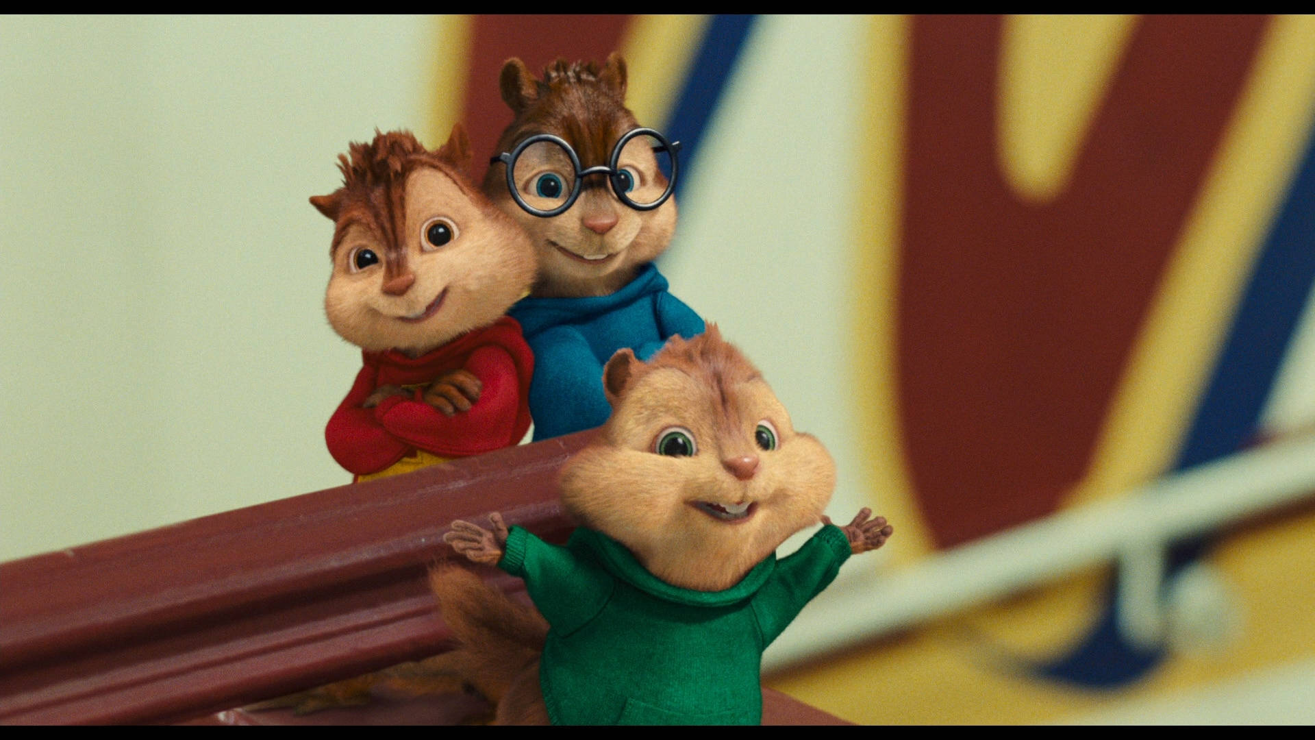 Chipmunks 桌布 壁紙 — 三隻動畫花栗鼠穿著色彩繽紛的毛衣，在模糊的體育場背景下開心地擺姿勢在欄杆上。