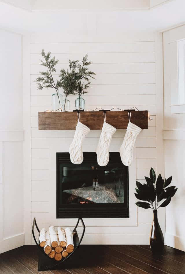 Christmas Living Room 桌布 壁紙 — 三隻奶油色針織長襪掛在一個質樸的木製壁爐架上，下方是一個點燃的壁爐，黑色支架上放著樺木柴。