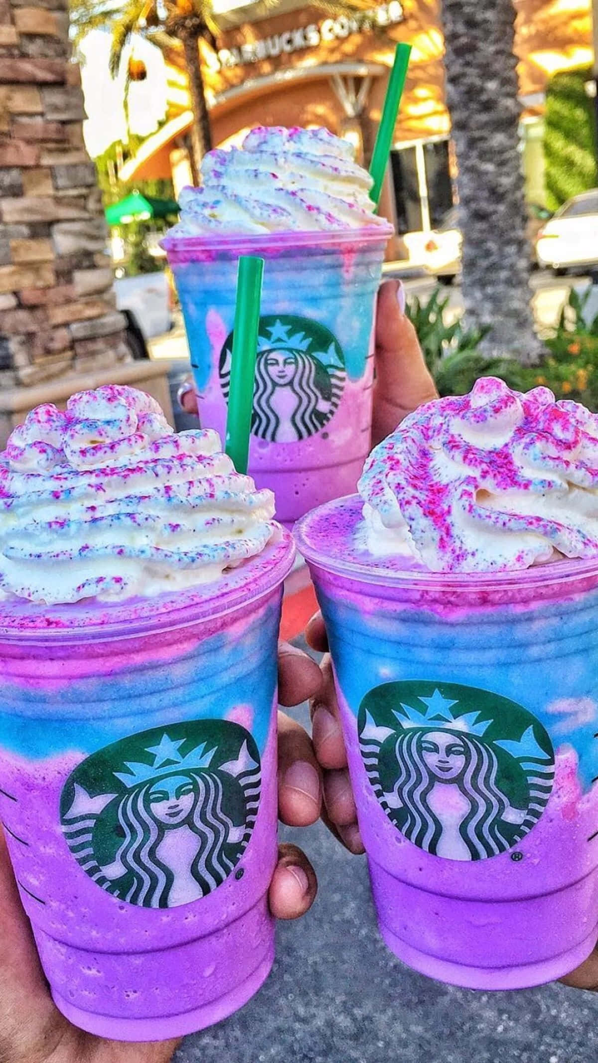 Aesthetic Starbucks 桌布 壁紙 — 三隻手在戶外舉著充滿活力的紫色和藍色星巴克星冰樂，上面有鮮奶油和粉紅色糖粒。