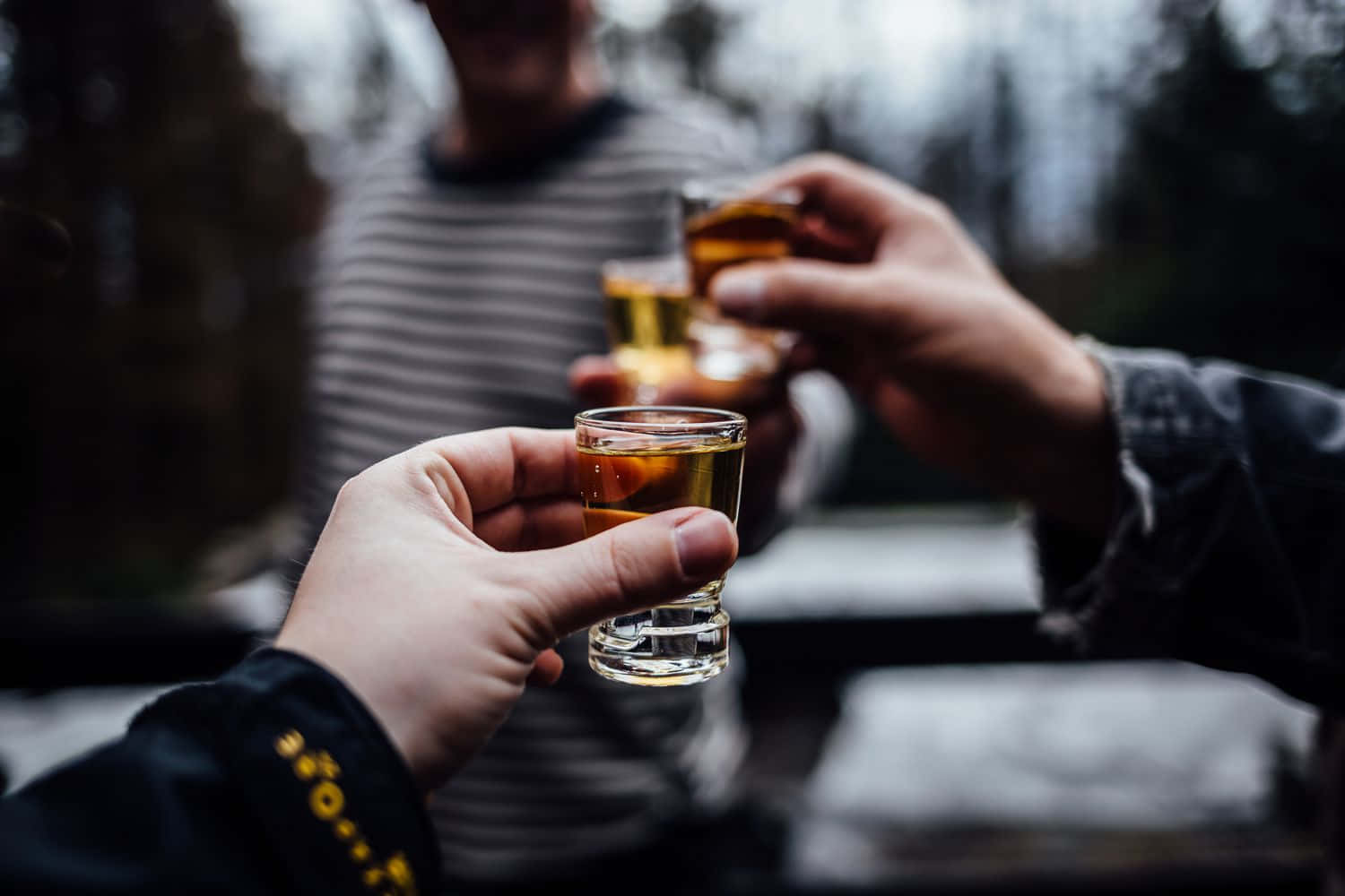醉意朦朧 桌布 壁紙 — 三隻手在模糊的戶外環境中碰杯，杯中裝滿琥珀色液體。