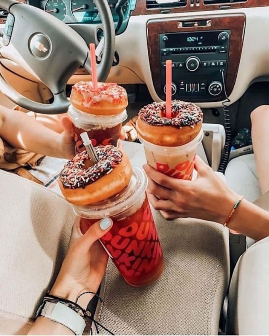 Dunkin Donuts 桌布 壁紙 — 三隻手在米色內裝和木紋飾邊的車內，拿著上面有甜甜圈的Dunkin' Donuts飲品。