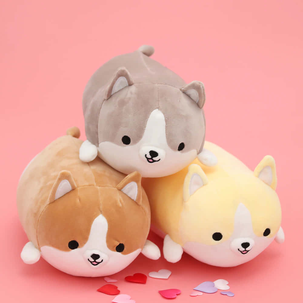 Kawaii Dog 桌布 壁紙 — 三隻灰色、棕色和黃色的胖乎乎柯基毛絨玩具放在一個散落心形的粉紅色背景上。