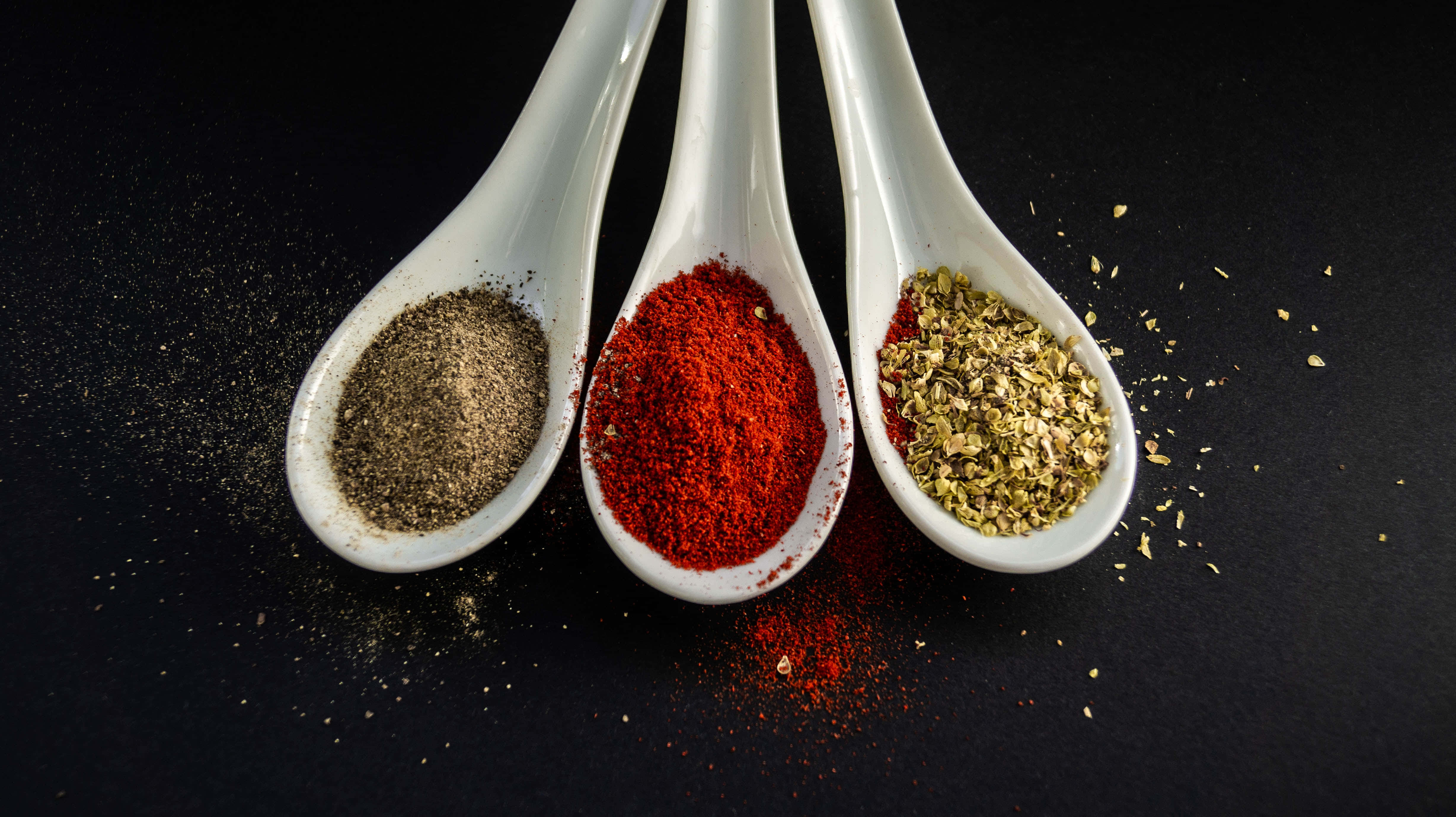 Spices 桌布 壁紙 — 三隻白色陶瓷湯匙盛載著鮮豔的香料，襯托著 stark 的黑色背景，並有散落的調味料。