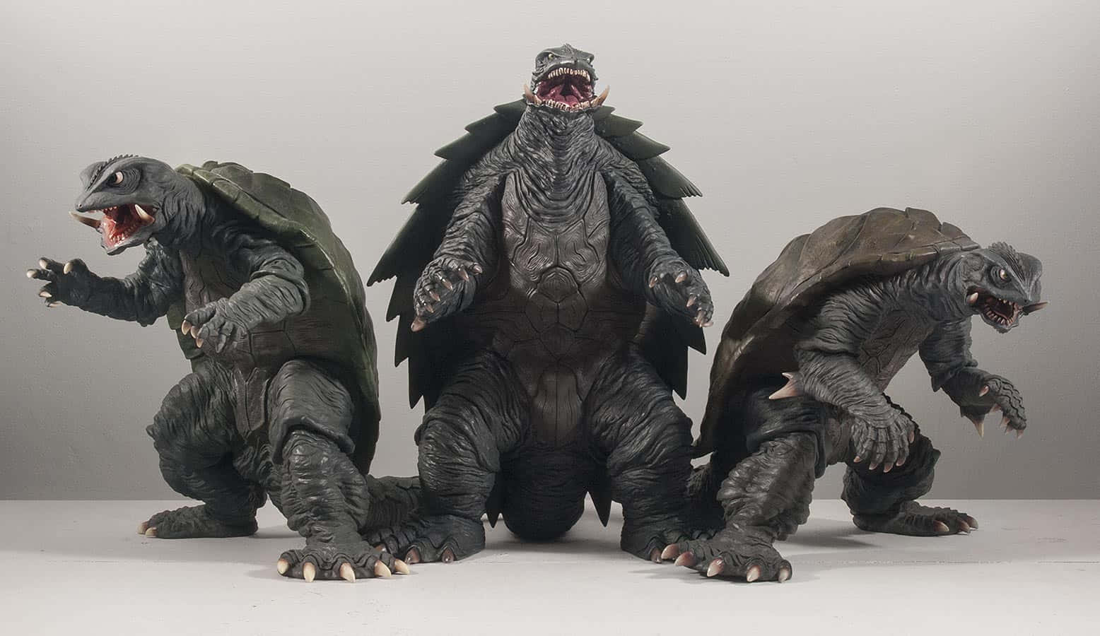 Gamera 桌布 壁紙 — 三隻細節豐富、帶有尖刺甲殼的哥斯拉風格烏龜怪物，在純灰色背景上兇猛咆哮。