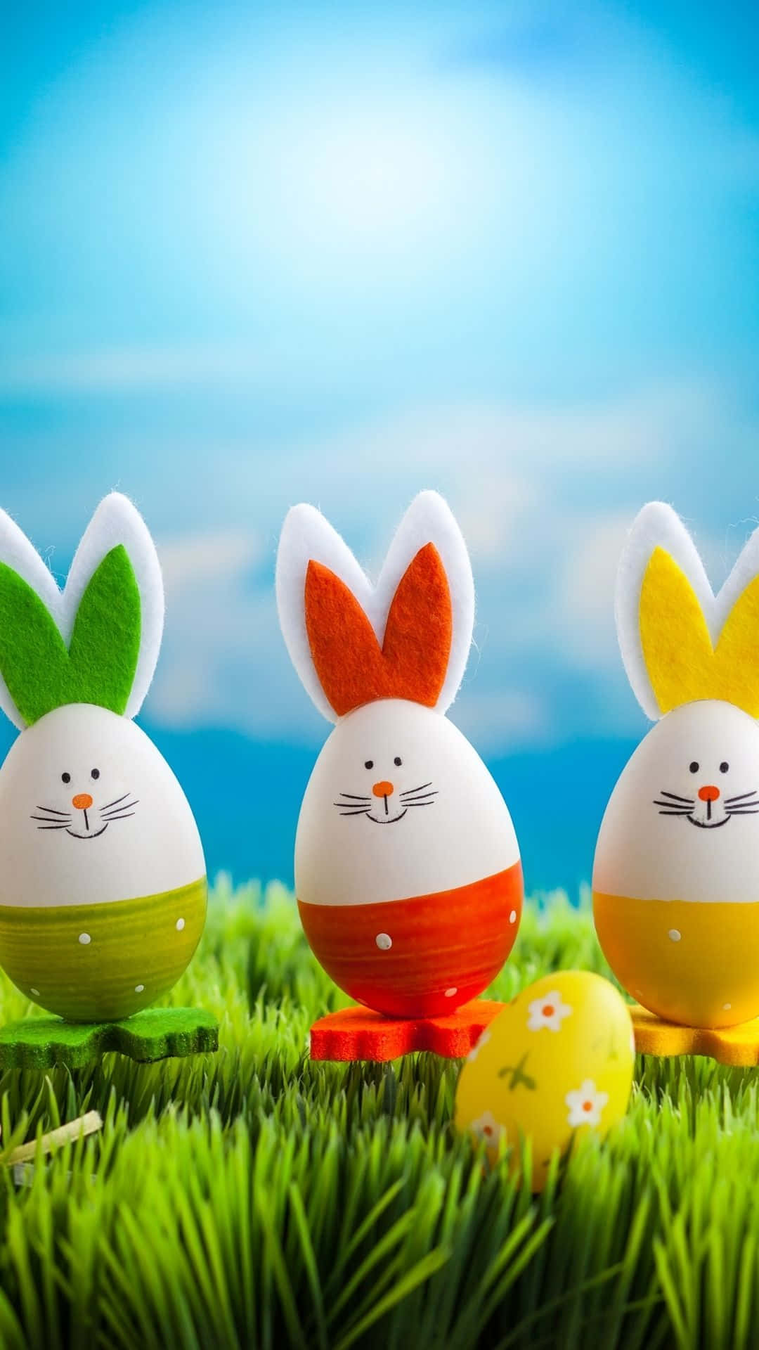 Easter Bunny 桌布 壁紙 — 三隻色彩繽紛的復活節蛋兔仔，帶有氈毛耳朵，站在充滿活力的綠草上，背景是明亮的藍天。