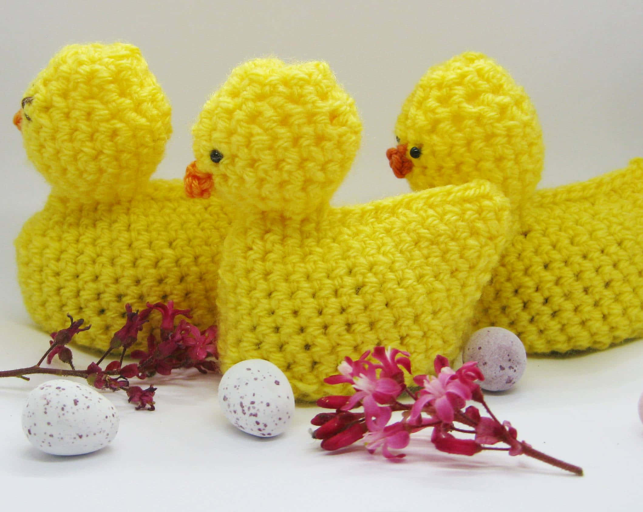 ducks 桌布 壁紙 — 三隻鮮黃色鉤針編織小雞坐在白色表面上，旁邊有斑點蛋和粉紅色花朵。