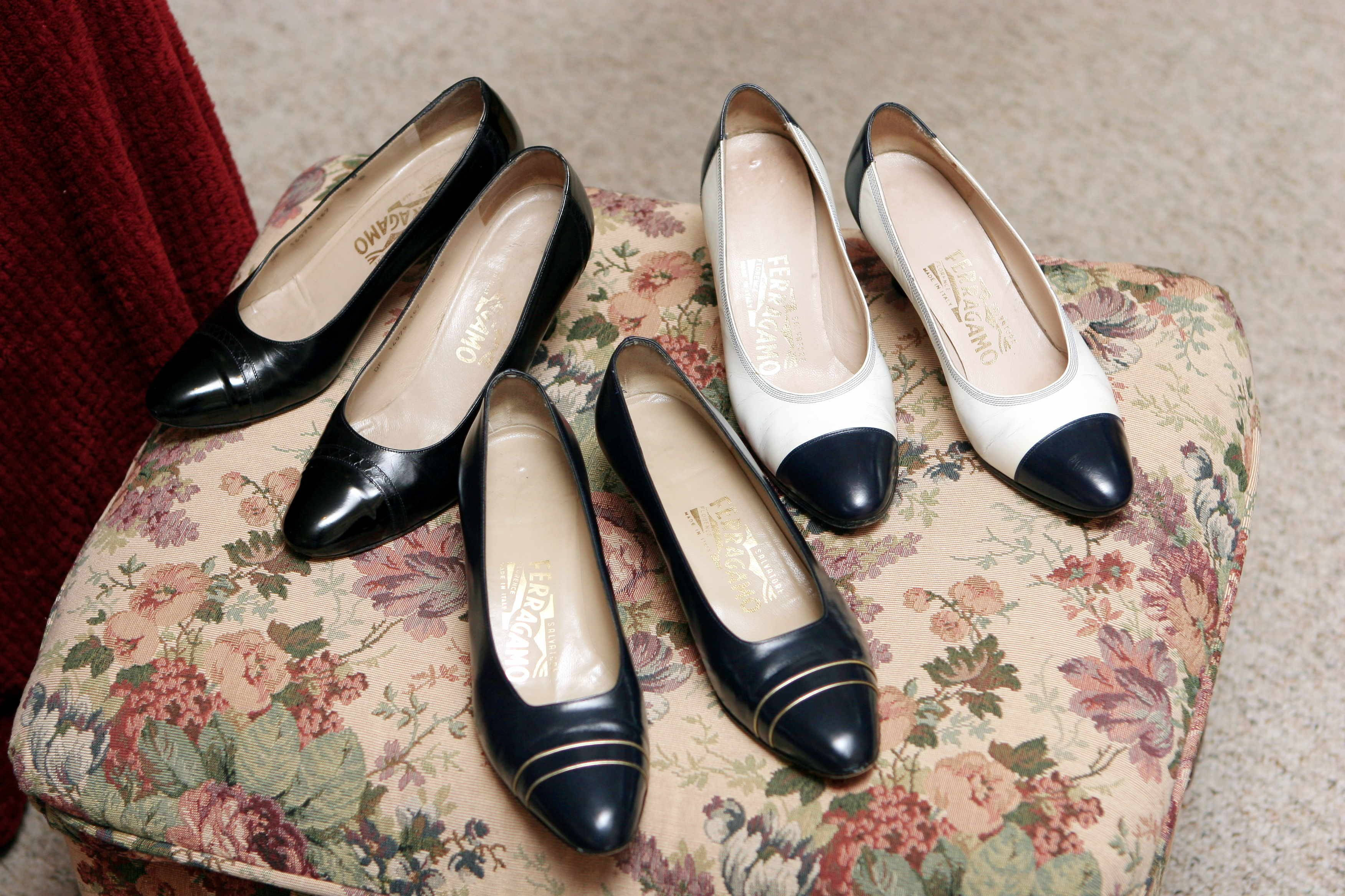 Ferragamo 桌布 壁紙 — 三雙優雅的女裝高跟鞋，黑色、白色和海軍藍色，放在一個復古花卉軟墊矮凳上。