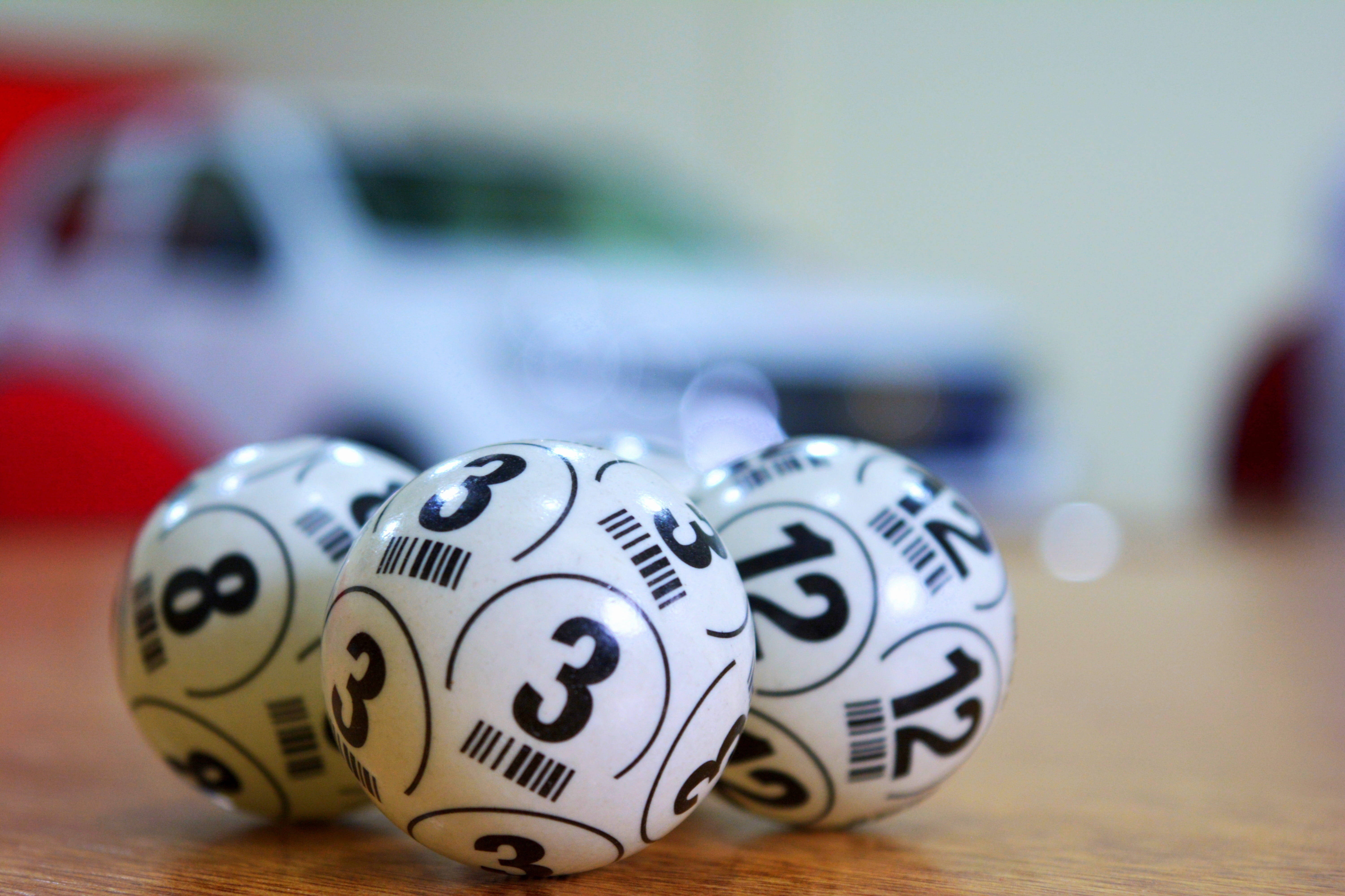 Powerball 桌布 壁紙 — 三顆光滑的彩券球，上面有數字三、八和十二，放在木質表面上，背景有一輛模糊的白色汽車。