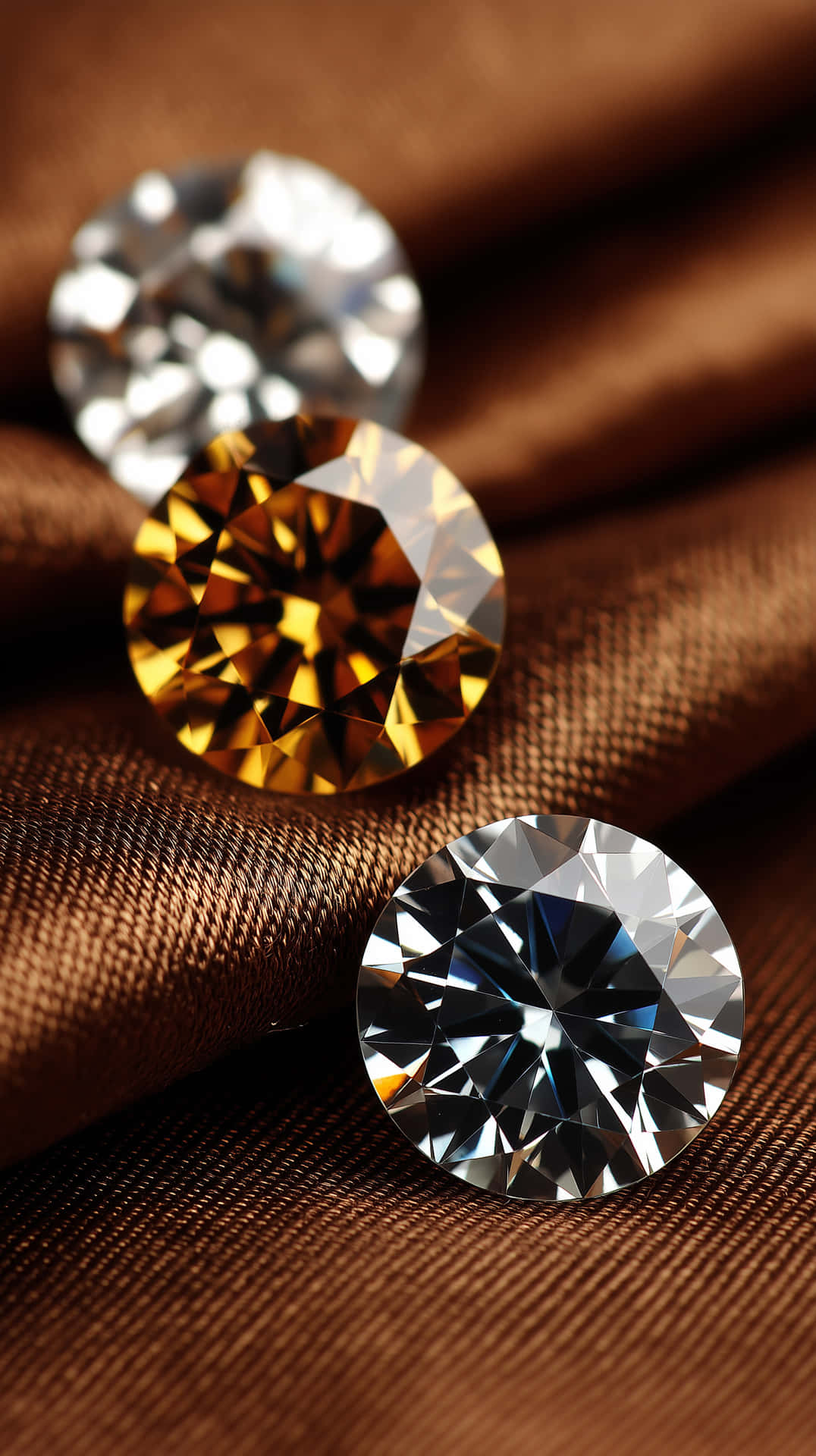 Silken Gem Assortment 壁紙 — 三顆圓形切割鑽石，一顆黃色、一顆透明、一顆藍色，放置在豐富的棕色布料上，帶有銳利的反射。