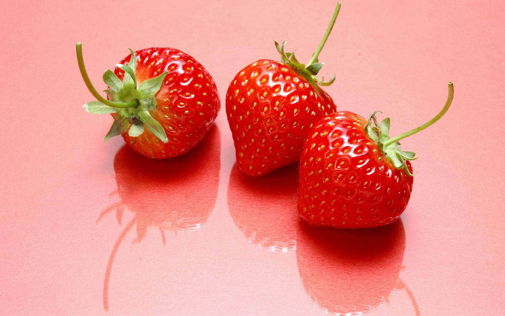 Red Strawberry 桌布 壁紙 — 三顆成熟的草莓，帶有綠色莖部，放置在光滑的粉紅色表面上，投射出柔和的倒影。