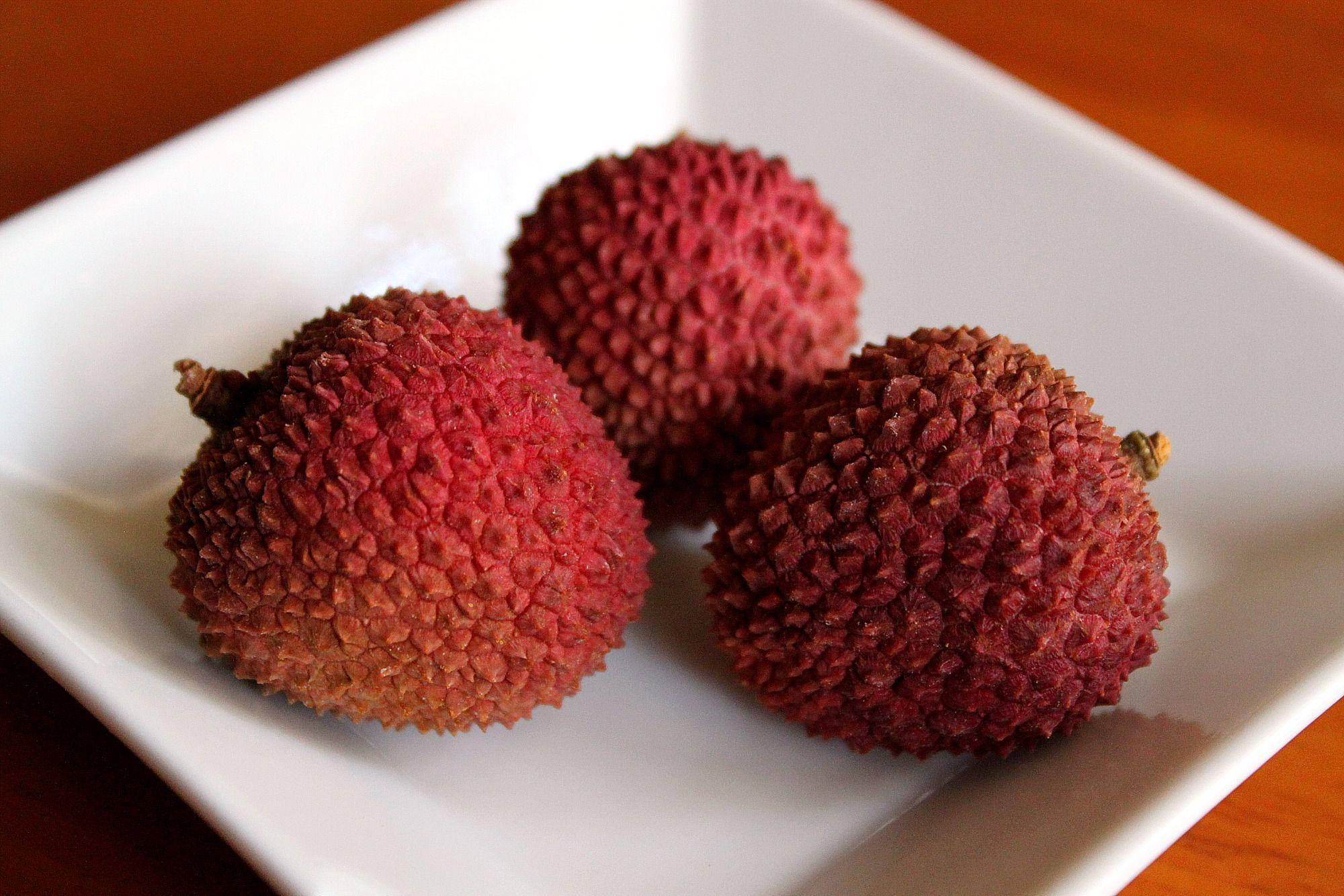 Litchi 桌布 壁紙 — 三顆成熟的荔枝，帶有紋理的紅色外皮，放在白色方形盤子上，背景為溫暖的木質。