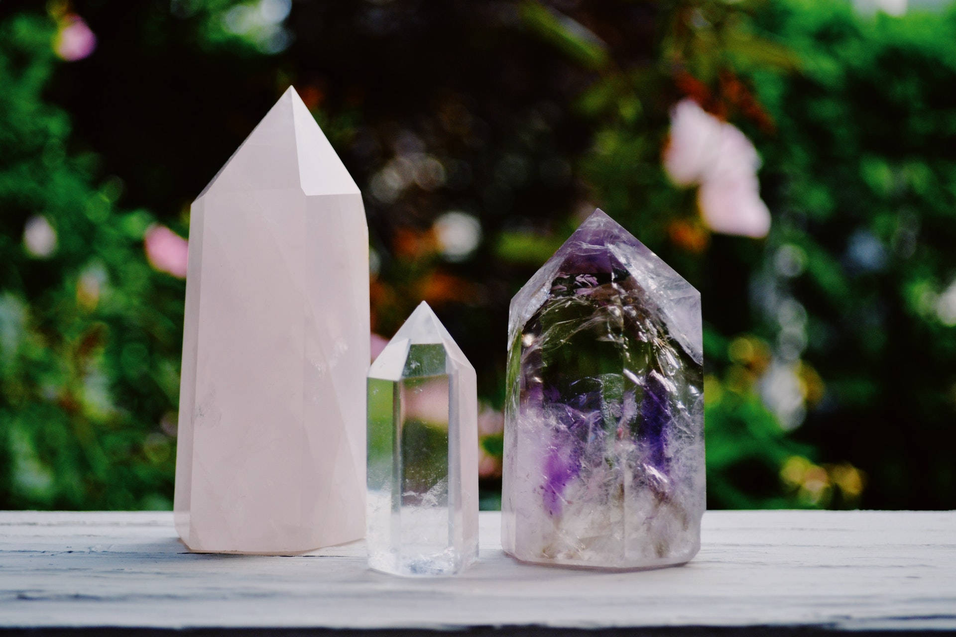 紫水晶 桌布 壁紙 — 三顆拋光的水晶尖 玫瑰石英 透明石英 紫水晶 放置在風化的木質表面上，背景是柔和模糊的花園。