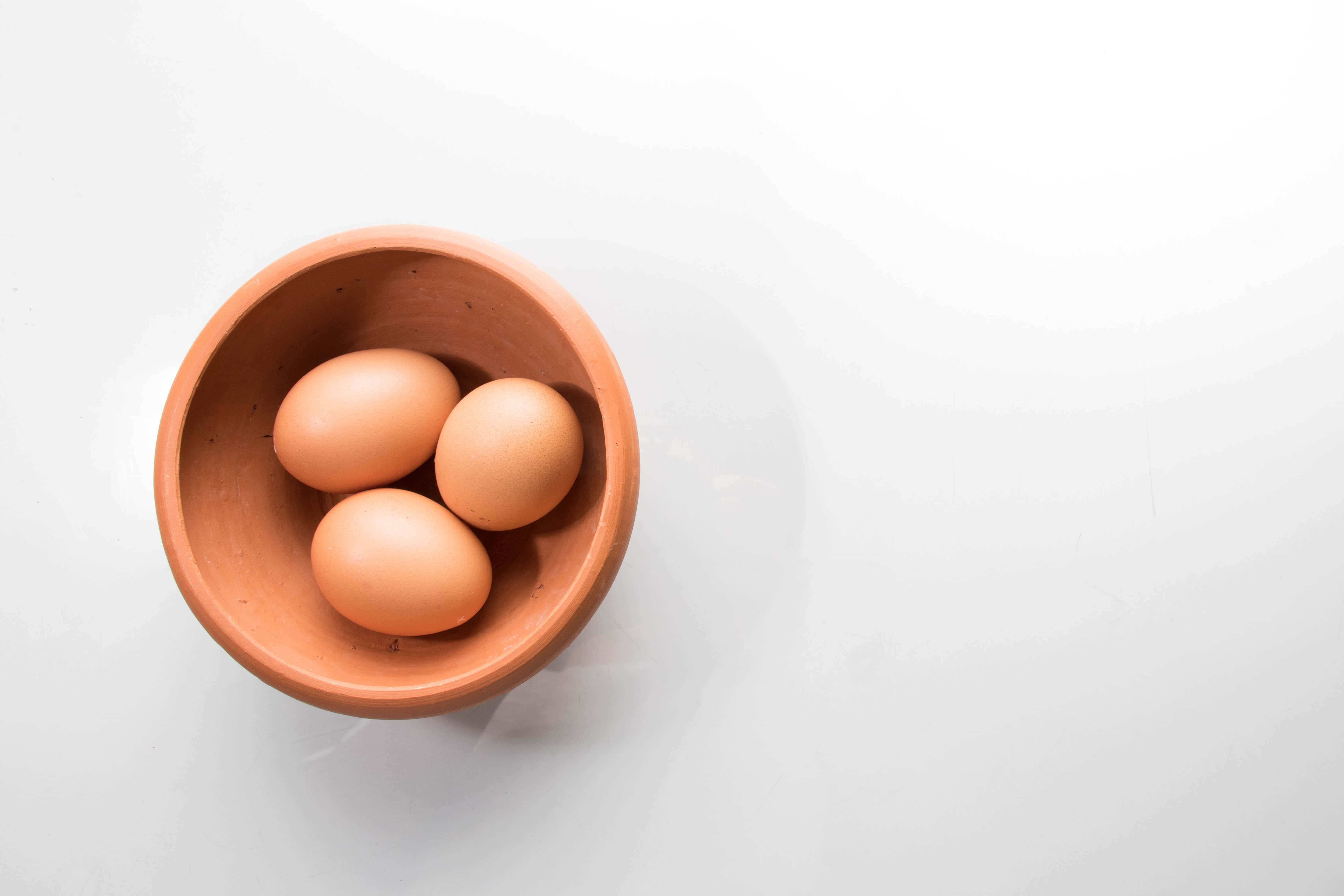 Brown Eggs 桌布 壁紙 — 三顆棕色雞蛋放在一個赤陶碗中，置於亮白色表面上，從正上方俯瞰。