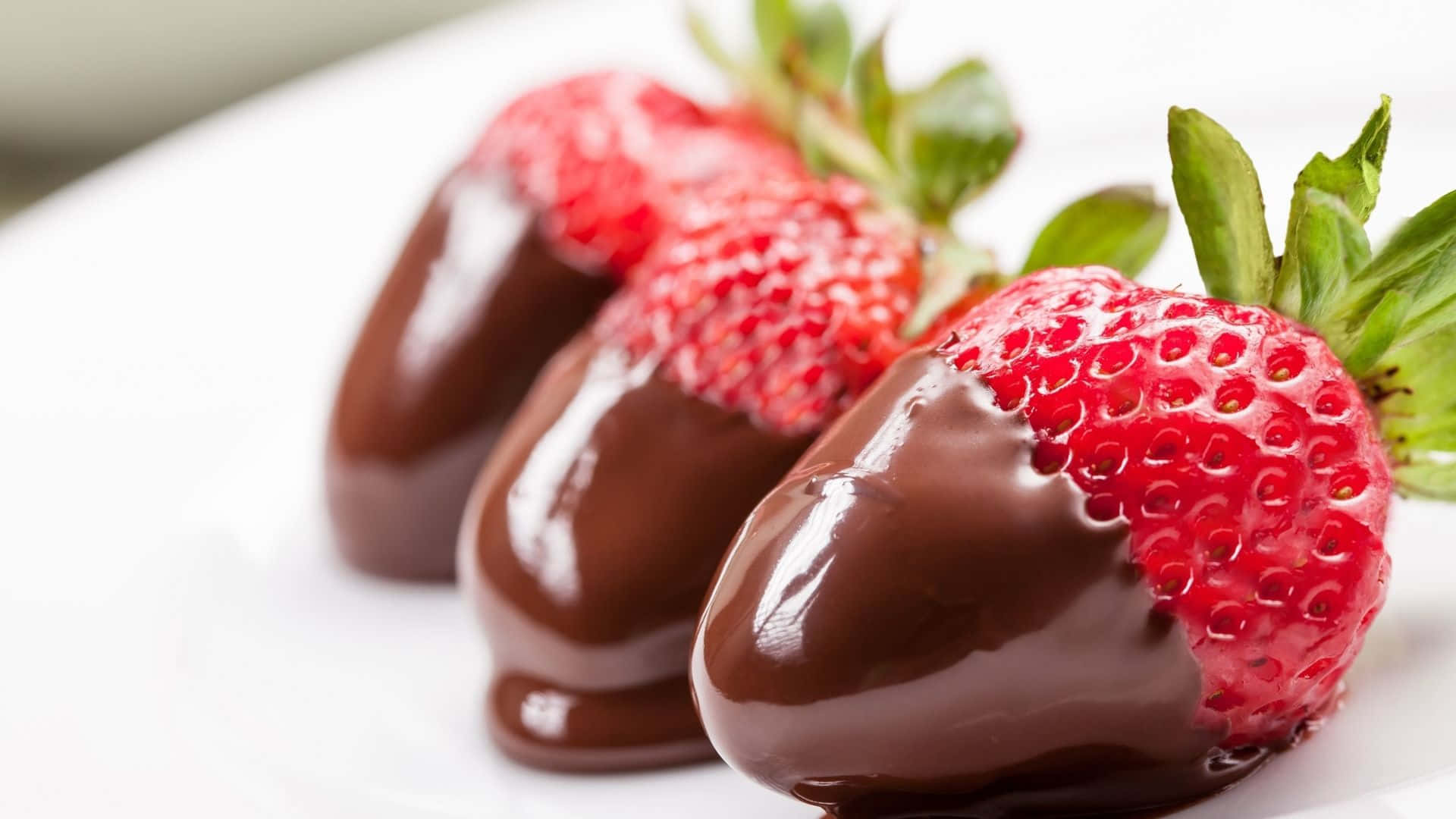 Red Strawberry 桌布 壁紙 — 三顆沾有巧克力的光澤草莓，帶有綠色頂部，柔和焦點下放在白色盤子上。