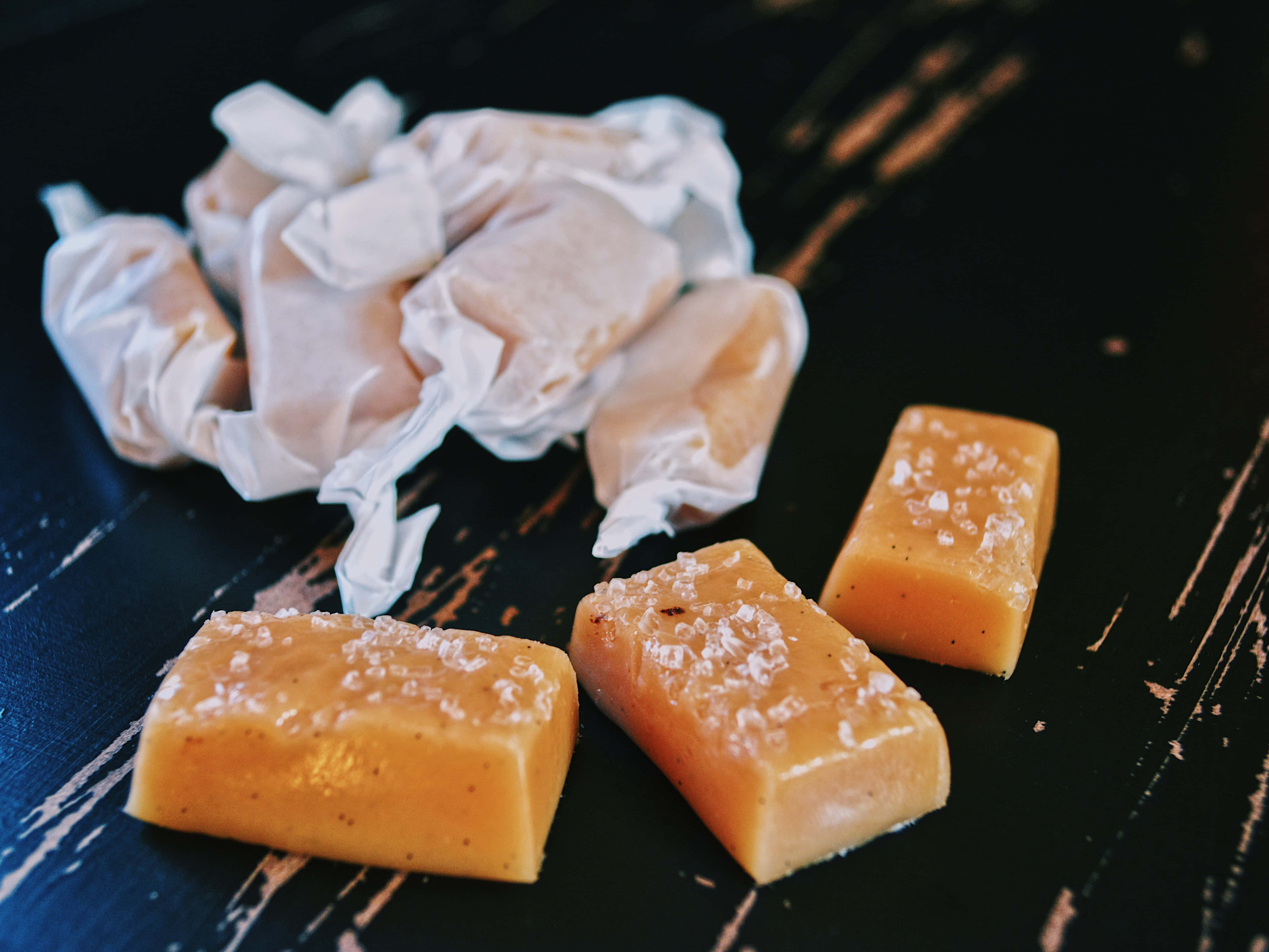 Caramel 桌布 壁紙 — 三顆海鹽焦糖糖果，帶有粗糖，放在深色鄉村風木面上，旁邊有皺巴巴的包裝紙。