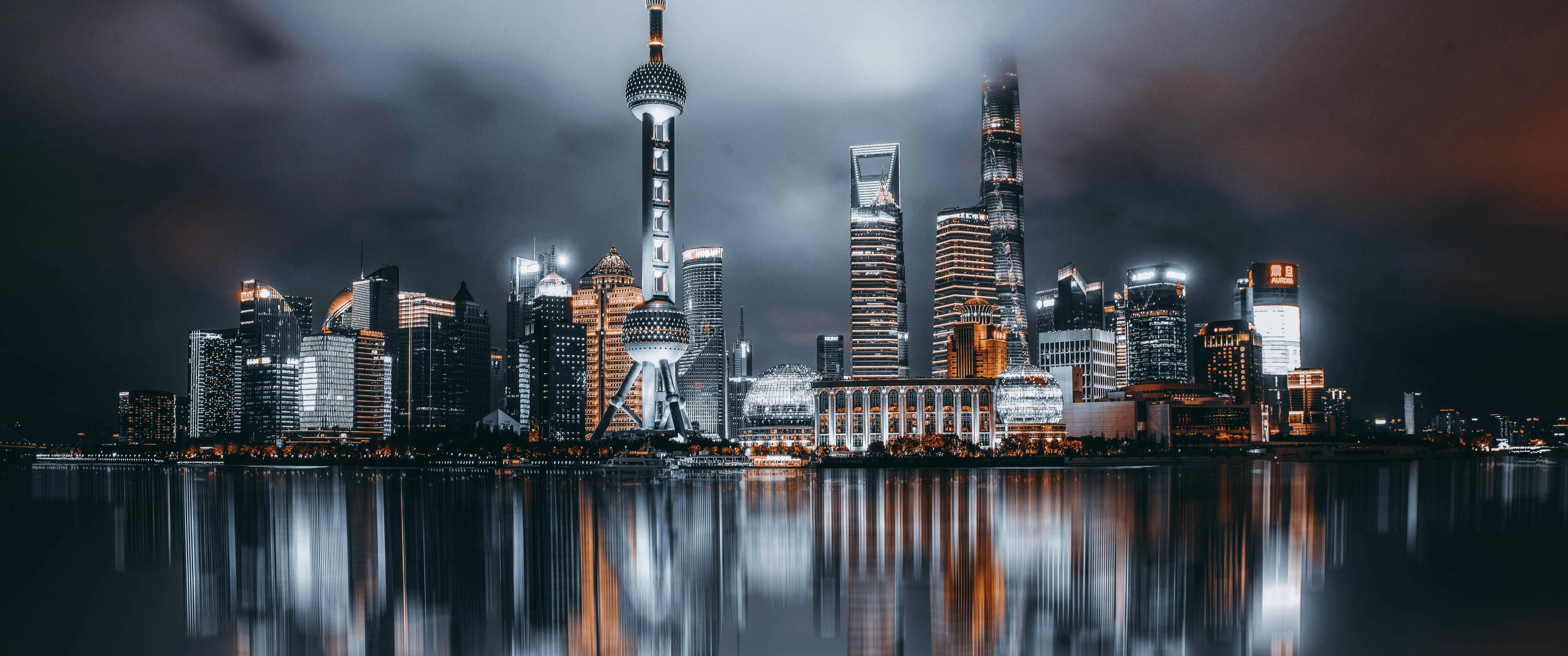 桌布 壁紙 — 上海天際線的戲劇性夜景，雲層密布的天空下，燈火通明的摩天大樓倒映在漆黑的水面上。