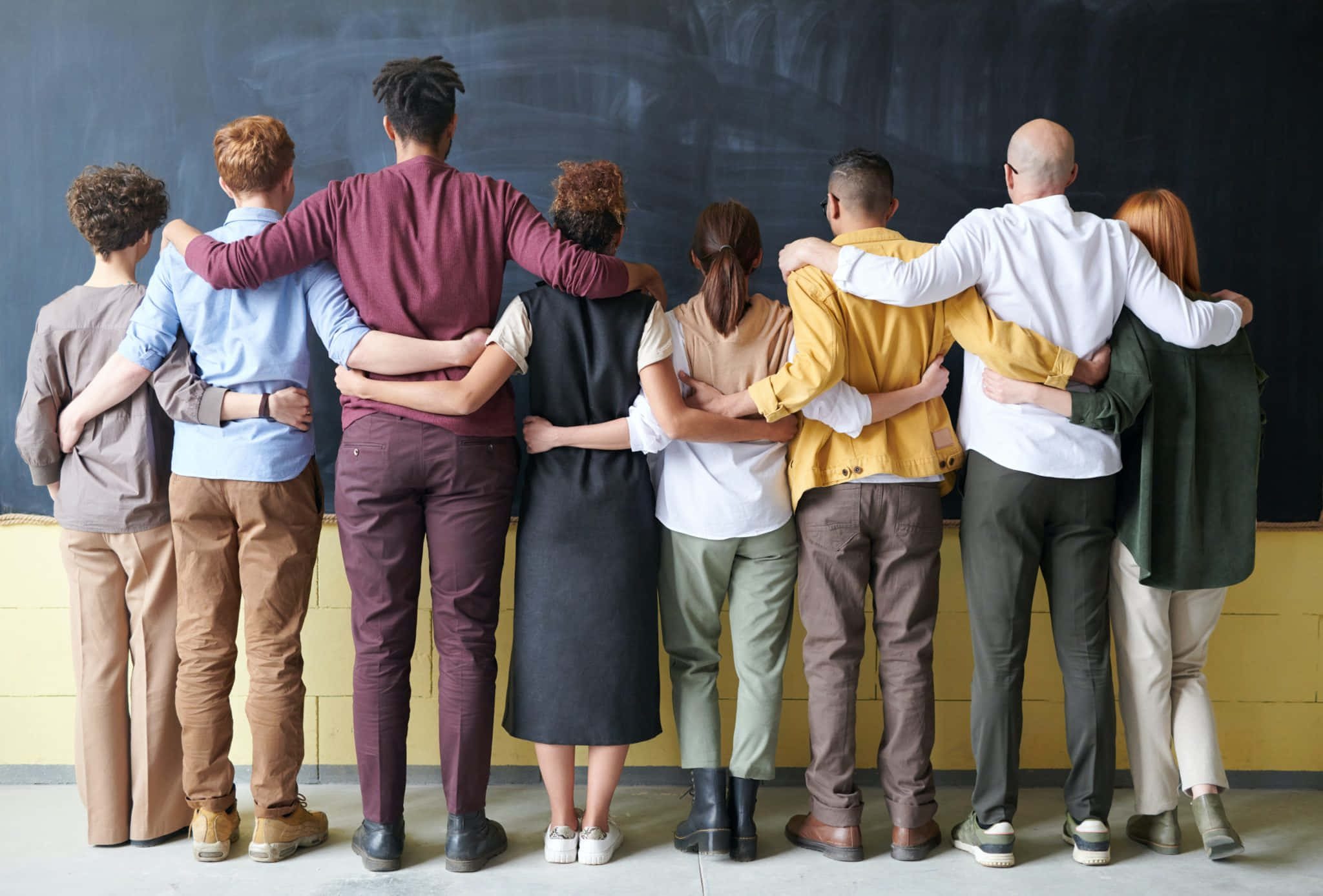 Students 桌布 壁紙 — 不同背景的人背對背站成一排，手臂搭在彼此的肩膀上，背景是黑板牆。