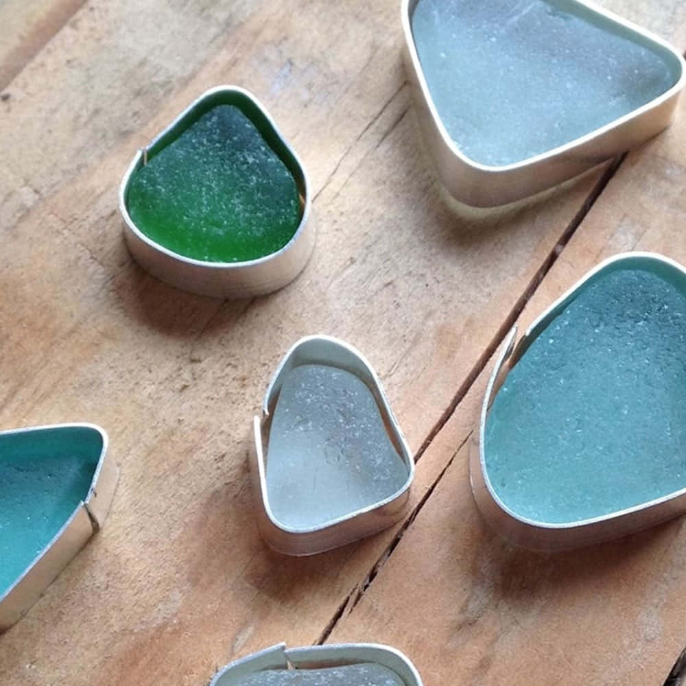 Sea Glass 桌布 壁紙 — 不規則形狀的海玻璃碎片，呈綠色和藍色色調，放在小金屬罐中，置於風化的木質表面上。