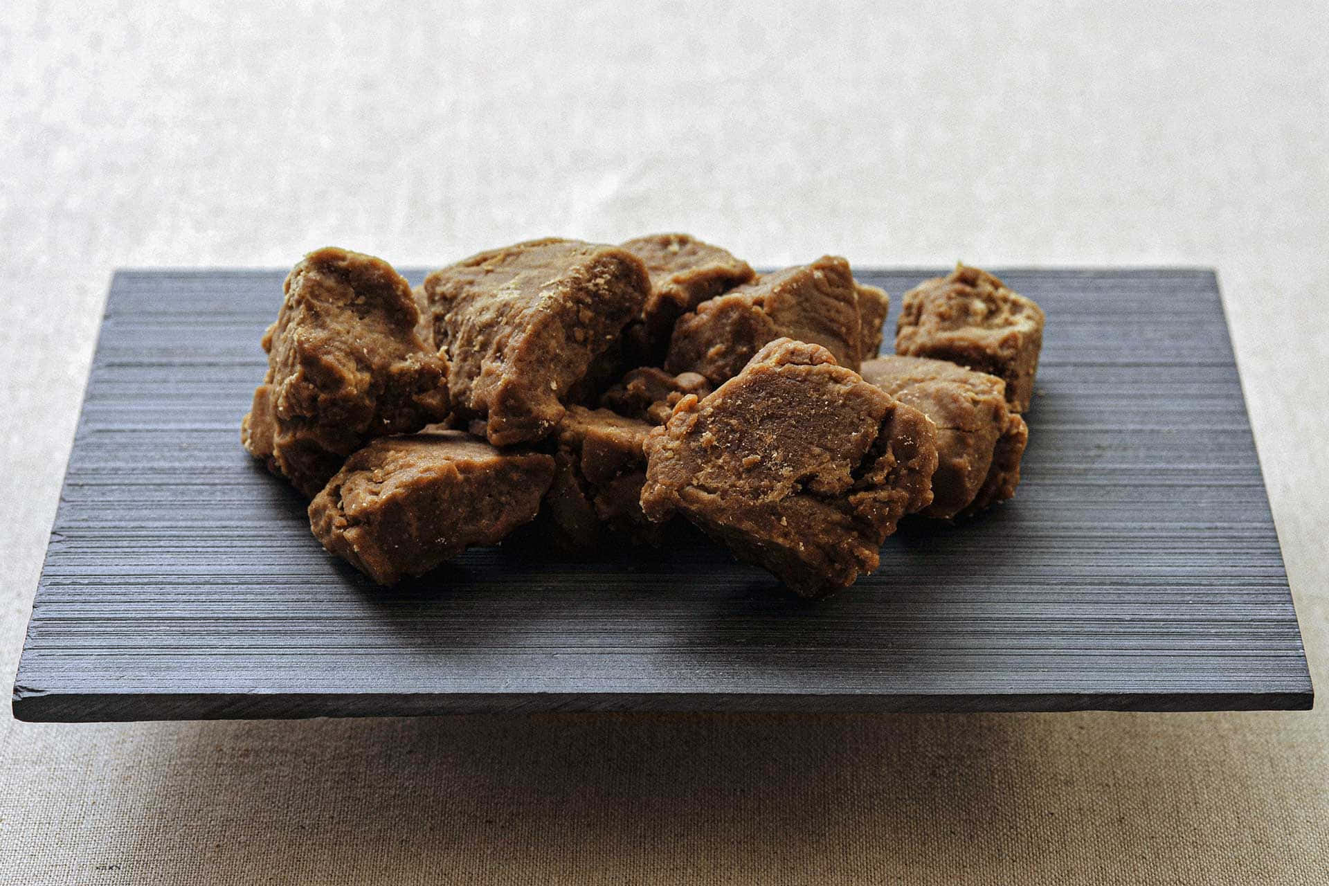Black Sugar 桌布 壁紙 — 不規則的棕色質地食物塊放在深灰色有棱紋的盤子上，背景是柔軟的米色布料。