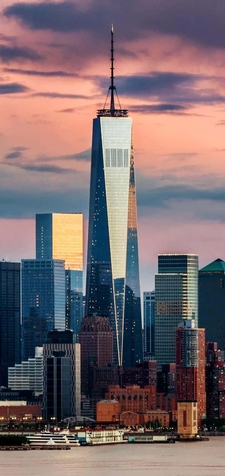 Skyscraper 桌布 壁紙 — 世界貿易中心一號大樓在日落時分聳立於紐約天際線之上，粉紅色和紫色的雲彩映照在玻璃摩天大樓和水面上。